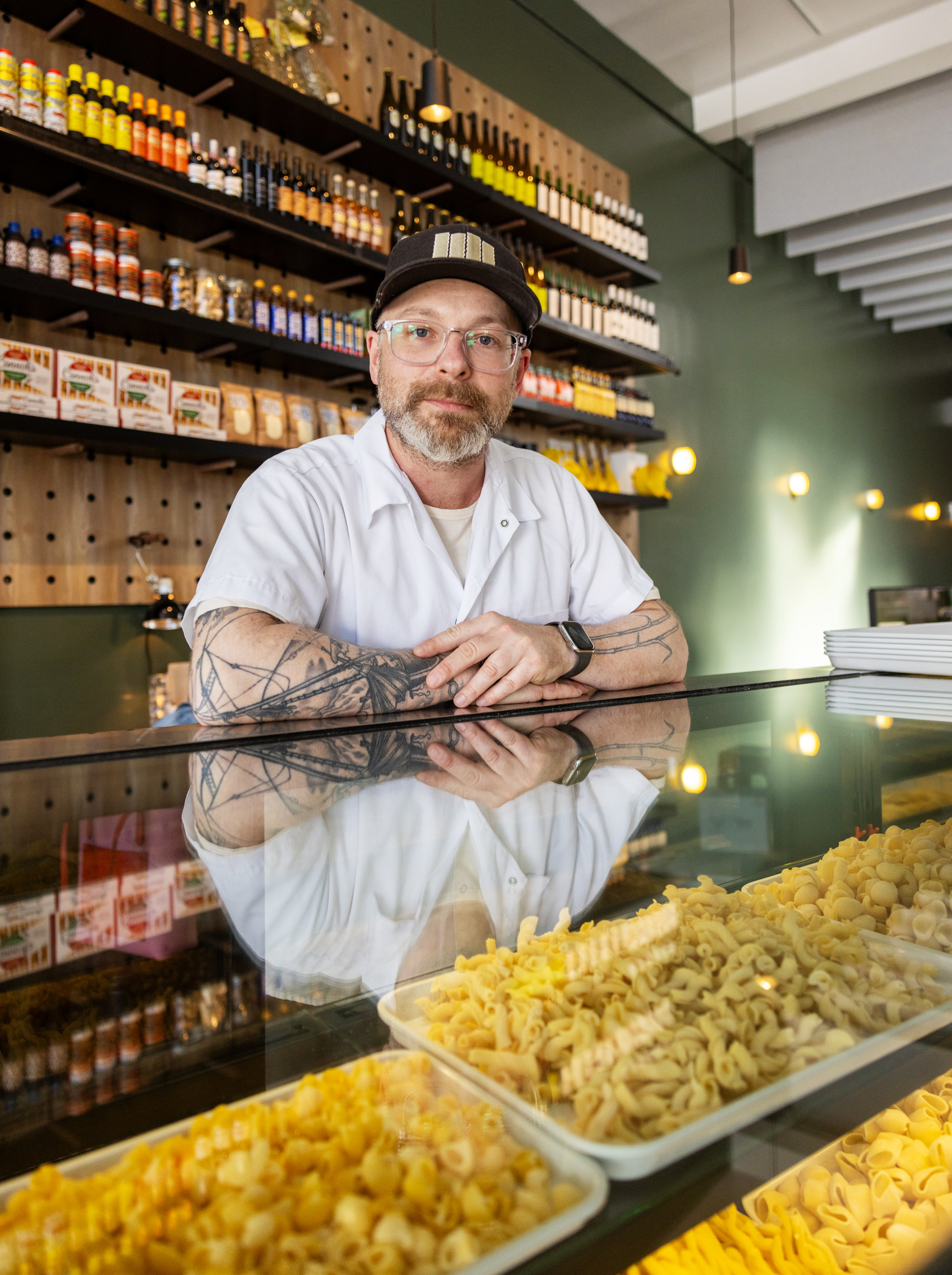 A bearded man wearing glasses and a hat stands behind a glass counter displaying different pasta types. Shelves with bottles and jars are behind him.