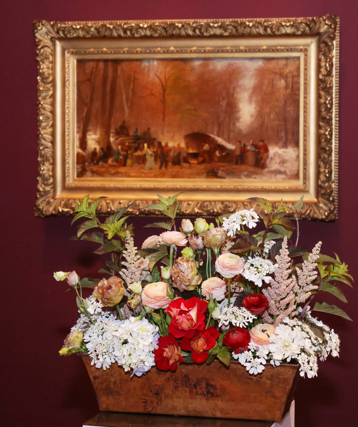 A gold-framed painting is hung on a maroon wall. Below it is a wooden planter with a colorful arrangement of flowers, featuring red, white, and pink blossoms.