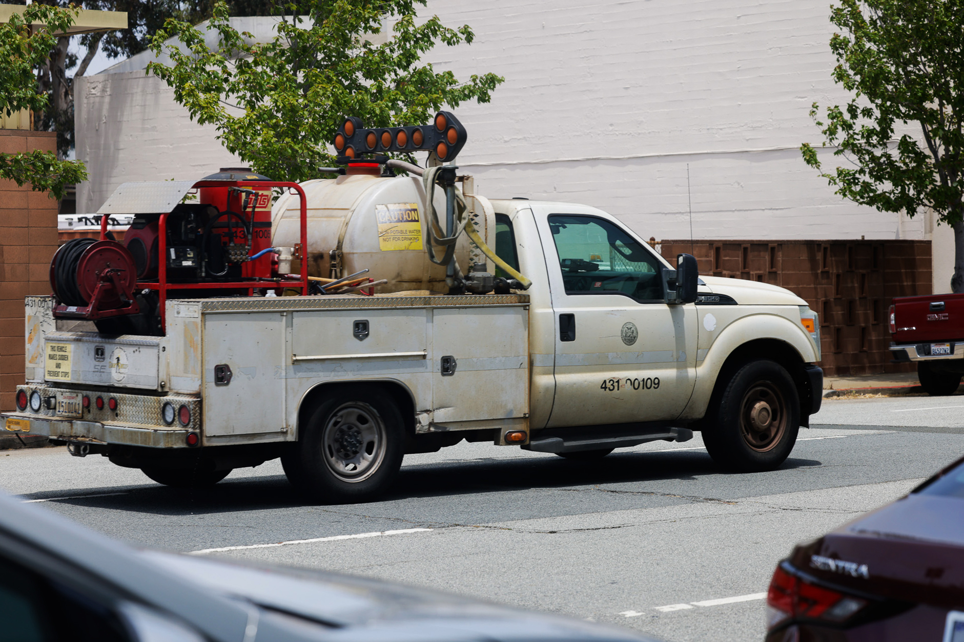 A utility pickup truck with equipment, including a large tank and hoses, driving on a city street.