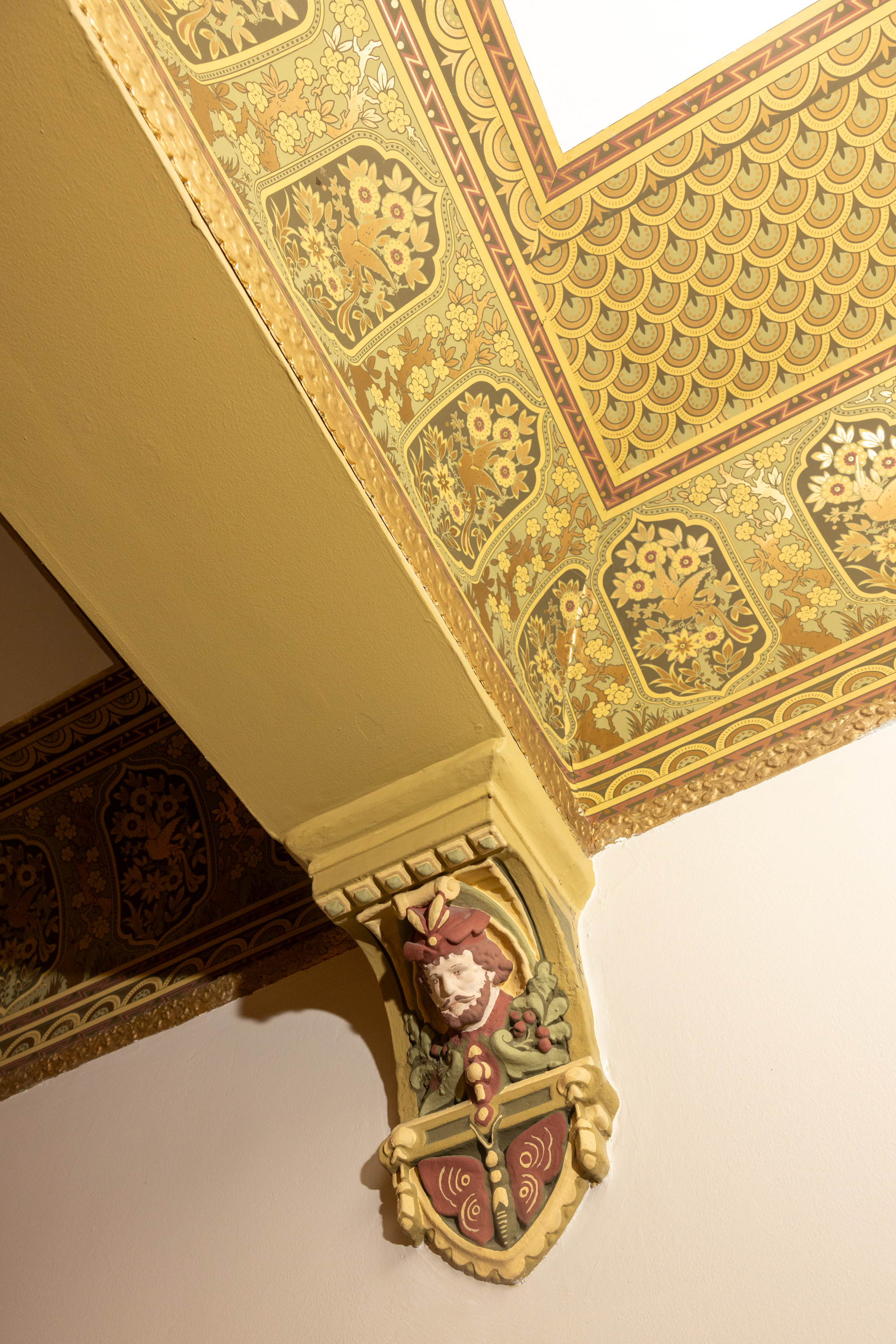 The image shows an ornate corner of a ceiling with intricate floral designs and a relief sculpture of a bearded man wearing a hat. The color palette is warm with gold and green tones.