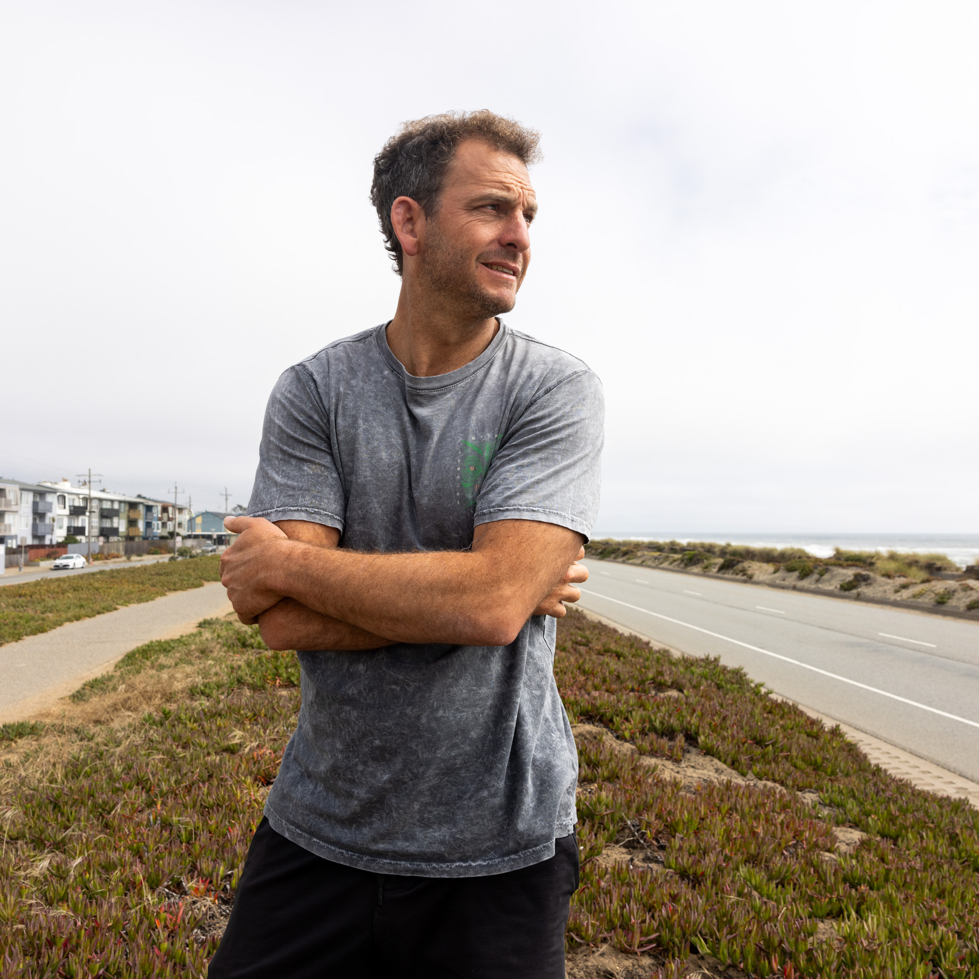 A man in a grey shirt stands with arms crossed, looking to his right. He is on a path near a road by the coast, with buildings and parked cars visible in the background.
