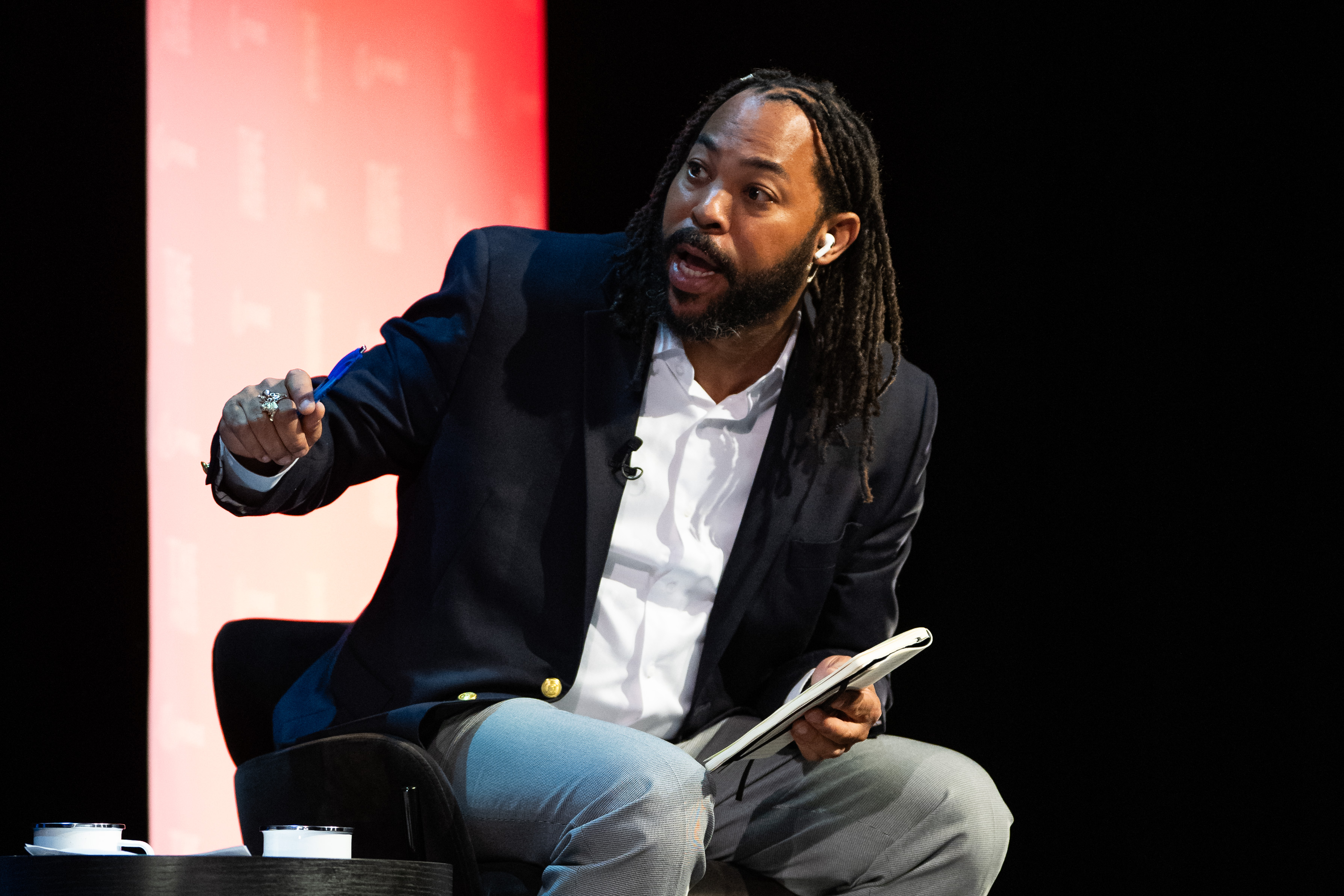 A man with dreadlocks, dressed in a dark blazer and light pants, is seated and holding a notebook. He is speaking animatedly, with a red and black backdrop behind him.