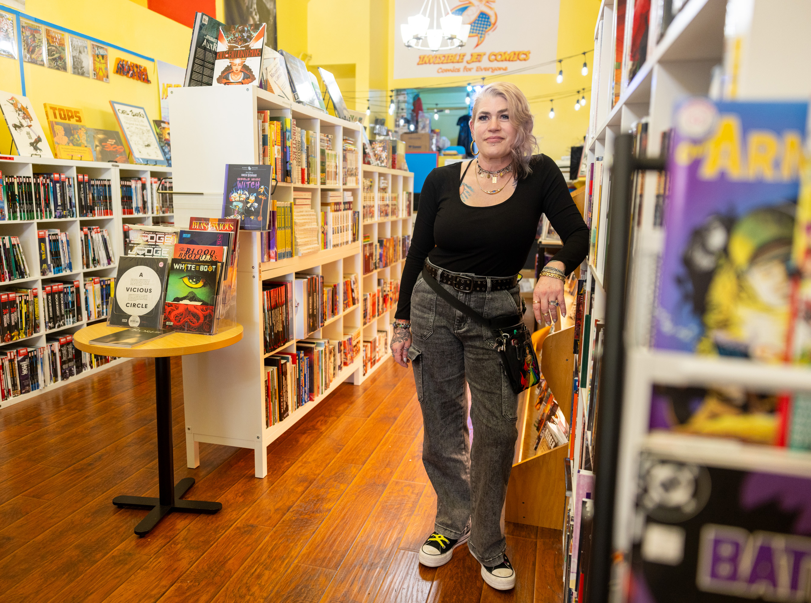 A person with light purple hair stands in a comic book store next to a shelf filled with books, with vibrant decor and organized displays around them.