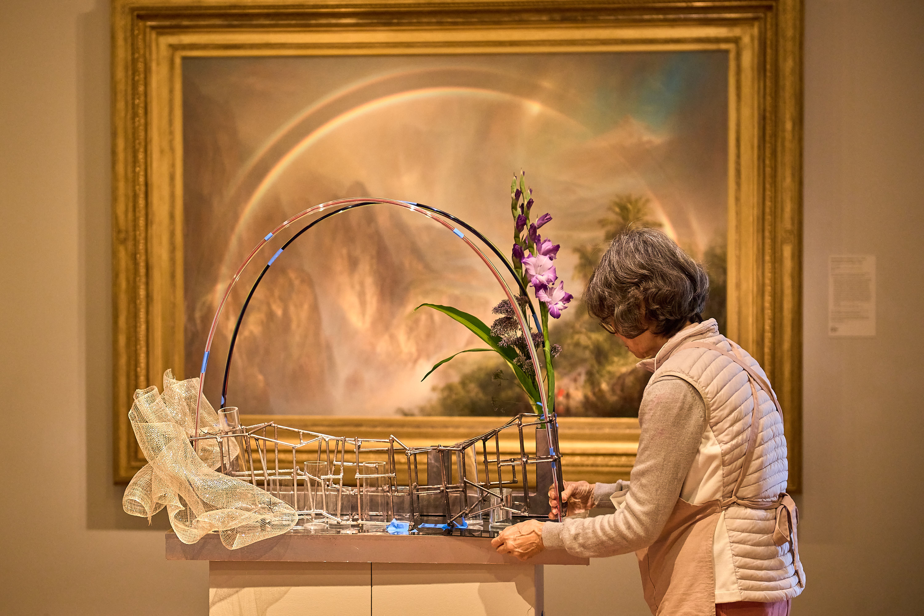 An older woman in a quilted vest arranges flowers in a metal sculpture. A vibrant painting with a rainbow and mountains is visible in the background.