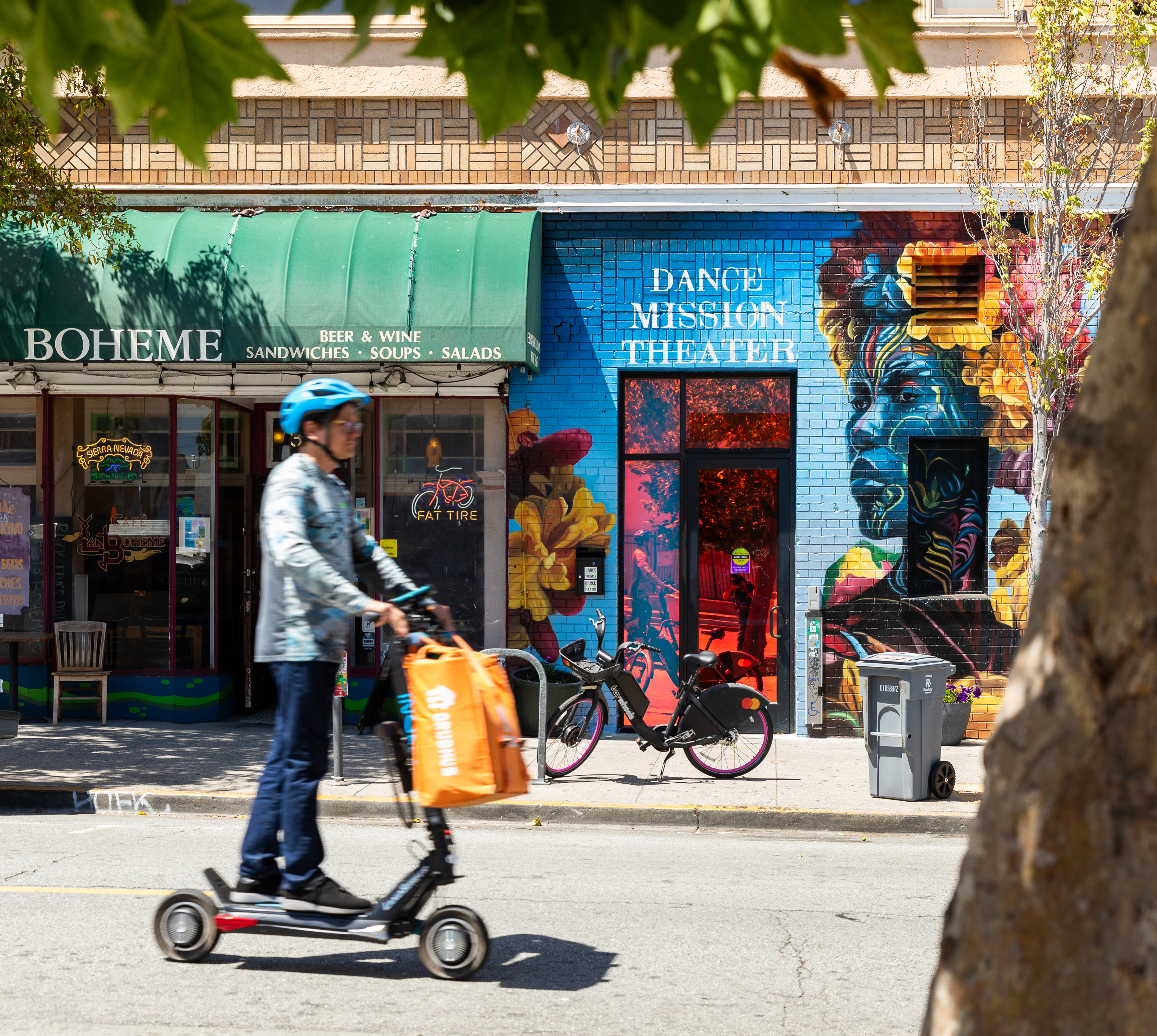 A person rides a scooter past a colorful mural and a shop named &quot;Boheme&quot; with a green awning offering sandwiches, soups, and salads.