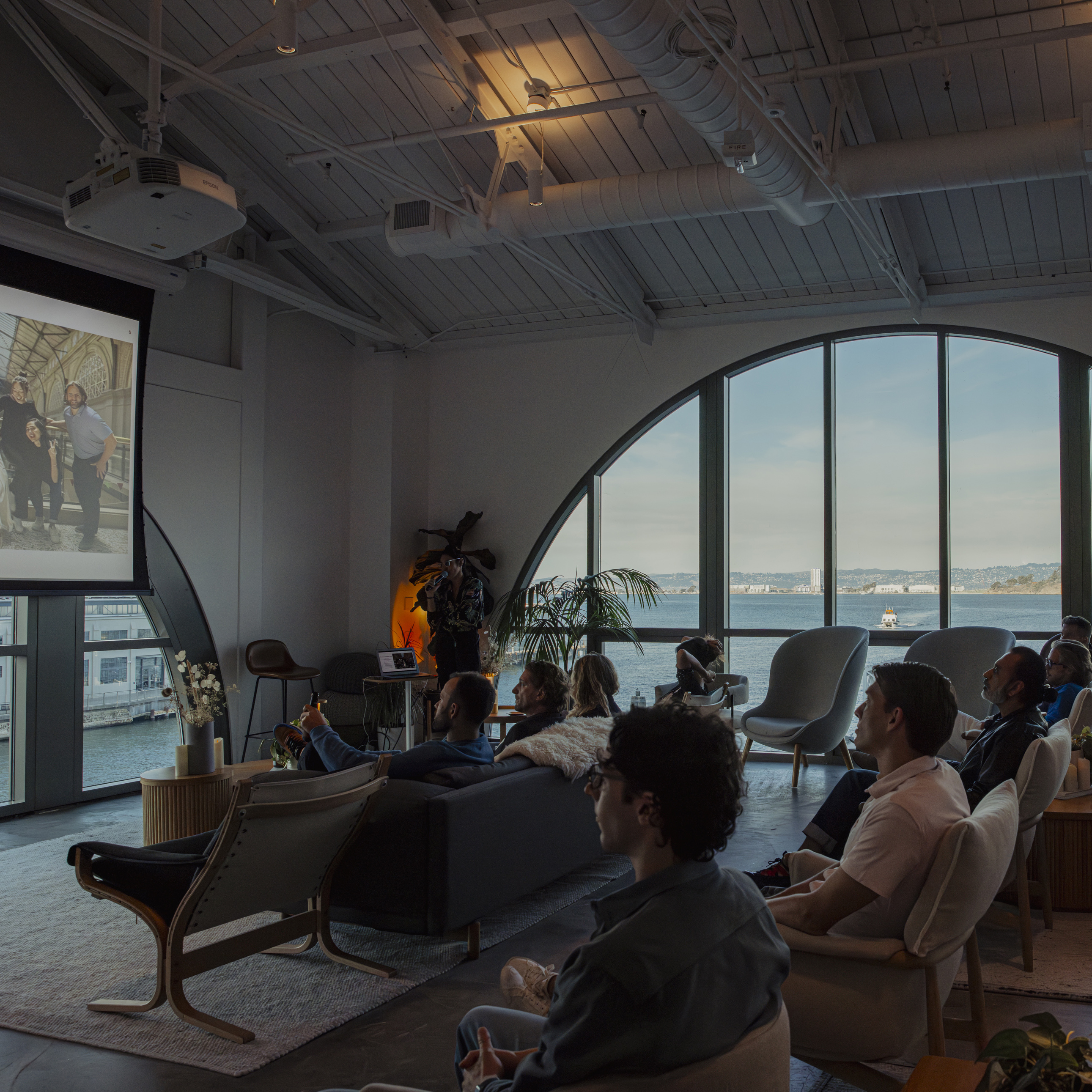 A group of people are seated in a modern room with large arched windows overlooking a body of water. They are watching a projected image on a screen.