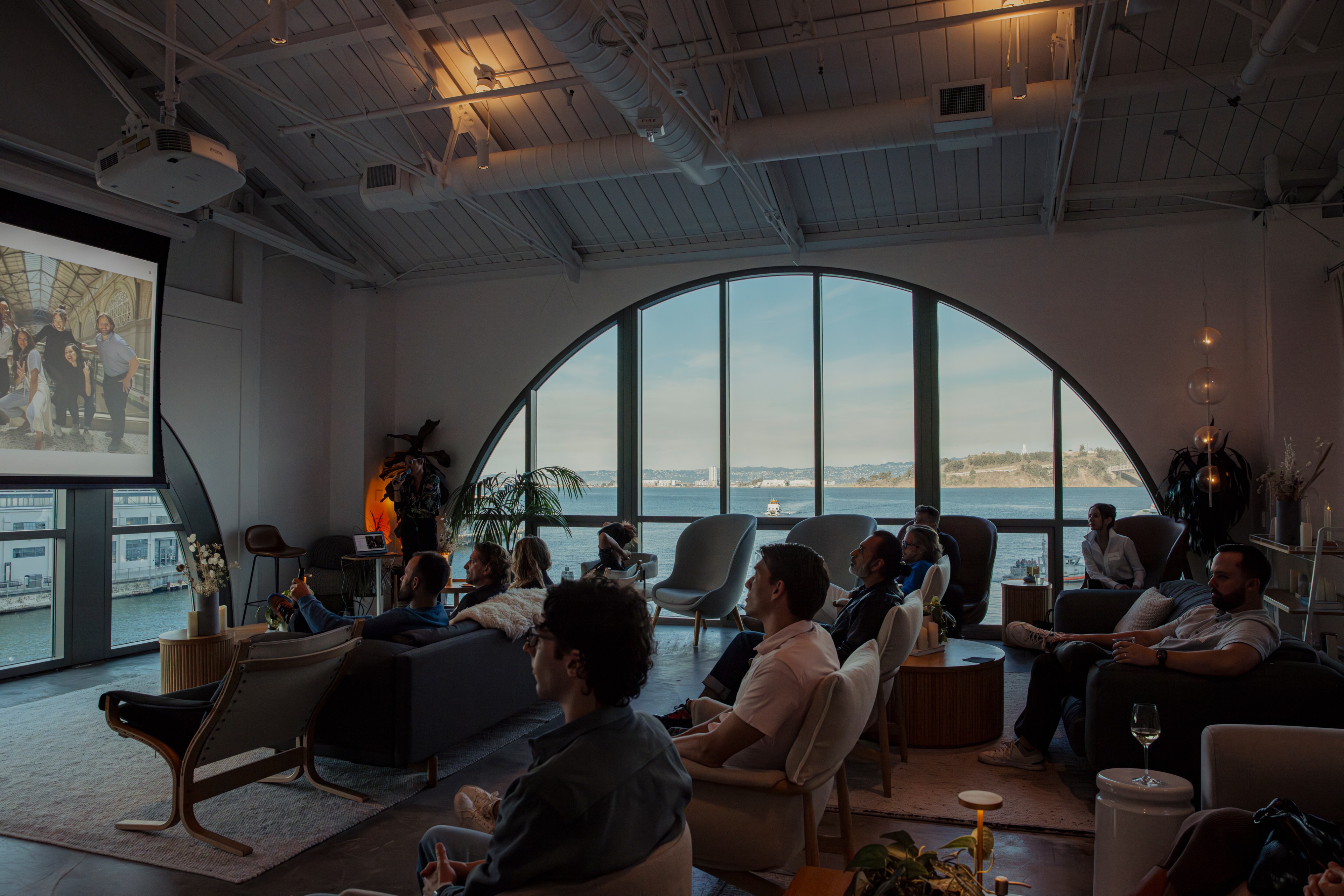 People are seated in a spacious room with large, arched windows overlooking a body of water. They are watching a presentation on a projected screen.