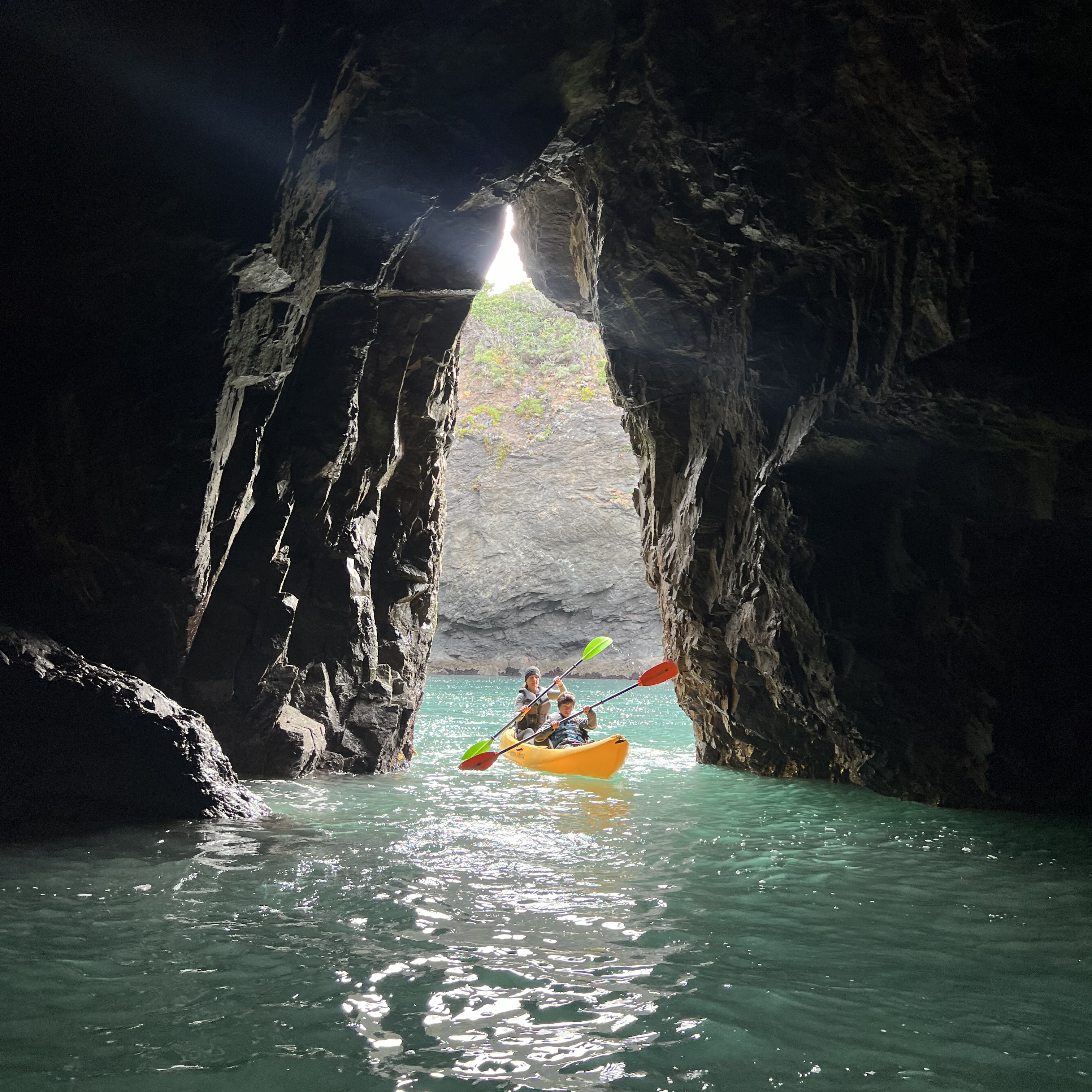 A kayak with two people paddles through the turquoise water inside a dark cave, heading toward a bright opening with rocky cliffs visible outside.