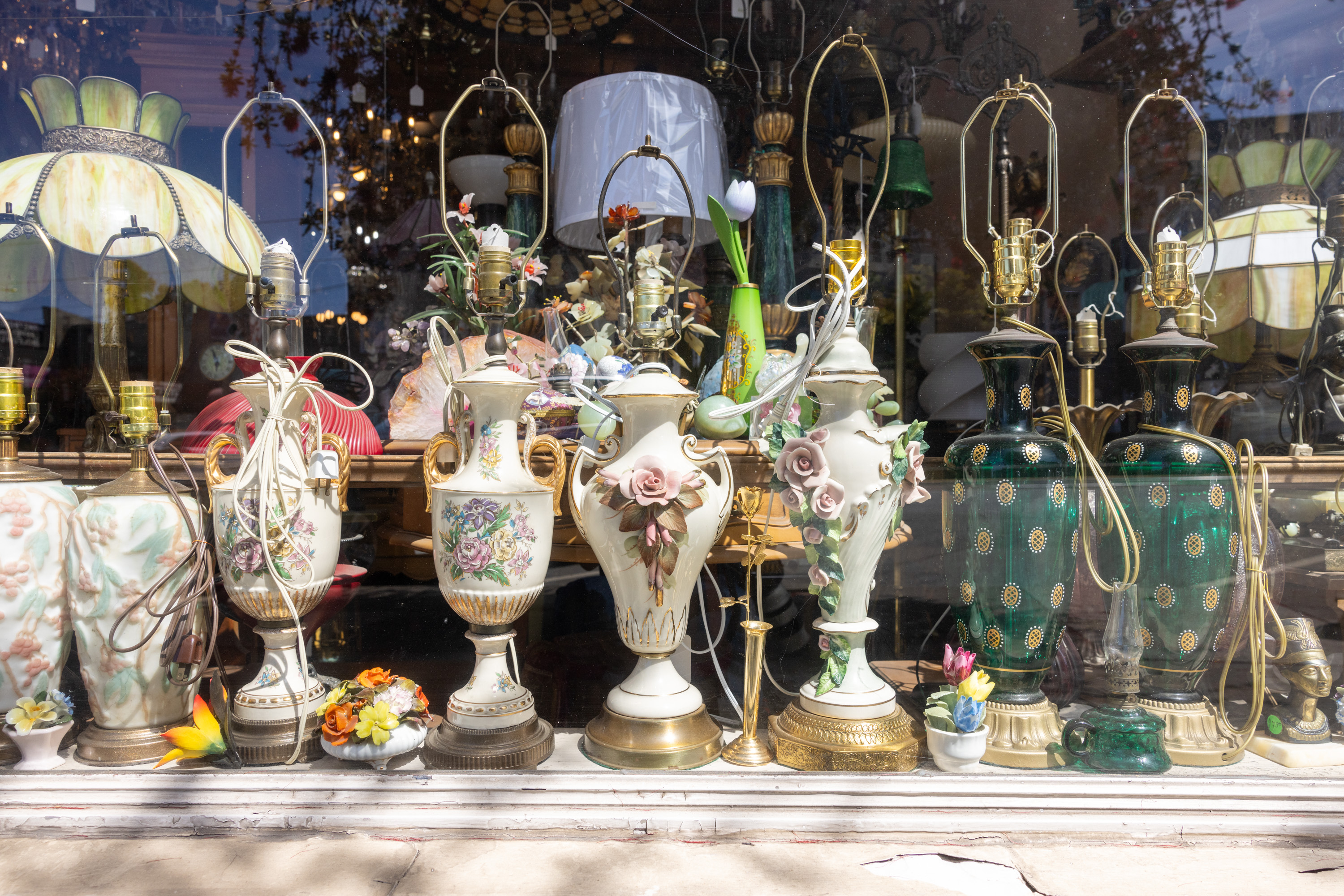 The image shows a shop window displaying an array of ornate, vintage-style table lamps and decorative vases, featuring intricate floral designs and vibrant colors.
