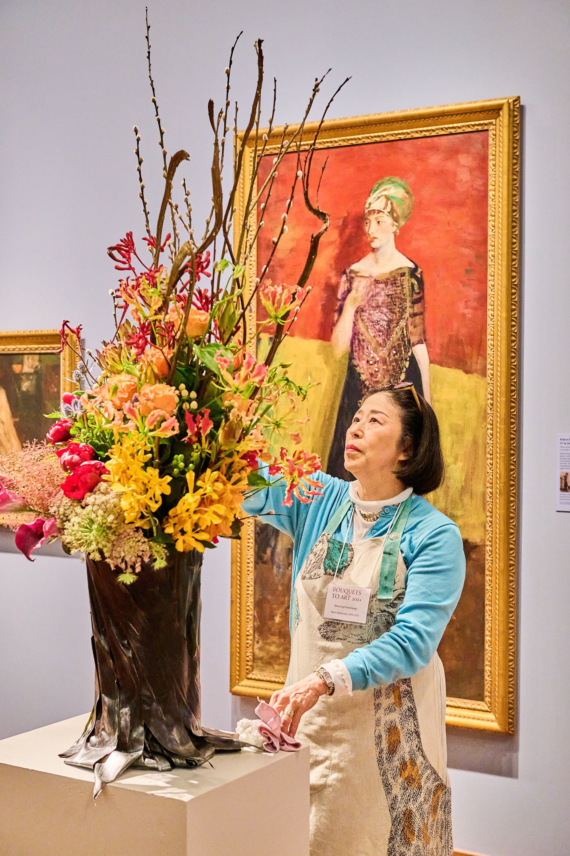A woman arranges a vibrant bouquet of flowers. She wears an apron and stands in front of a large framed painting of a woman in a red and yellow dress.
