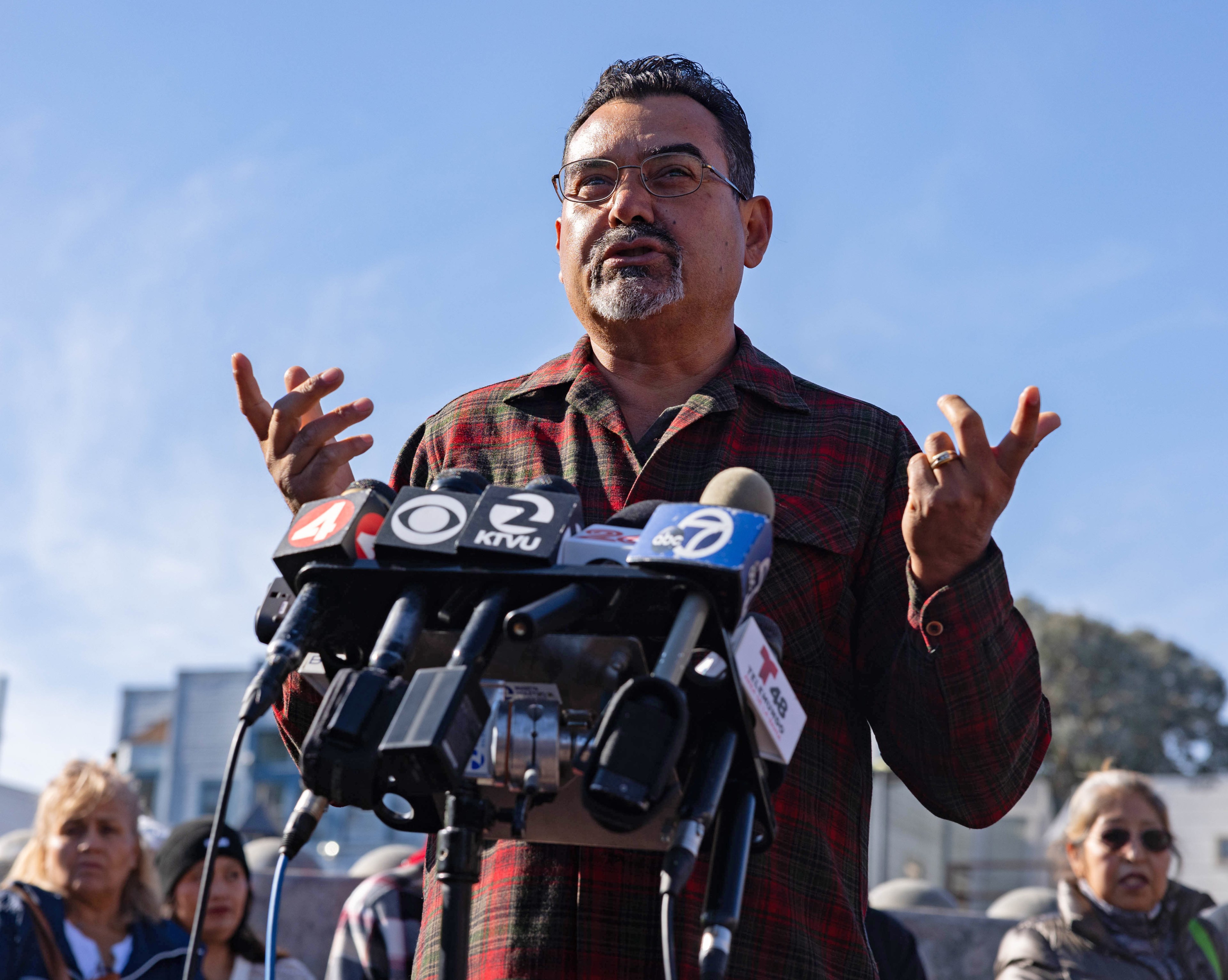 Rodrigo Lopez in a plaid shirt speaks passionately into multiple microphones at a press conference or public event, with people standing behind him against a clear blue sky.