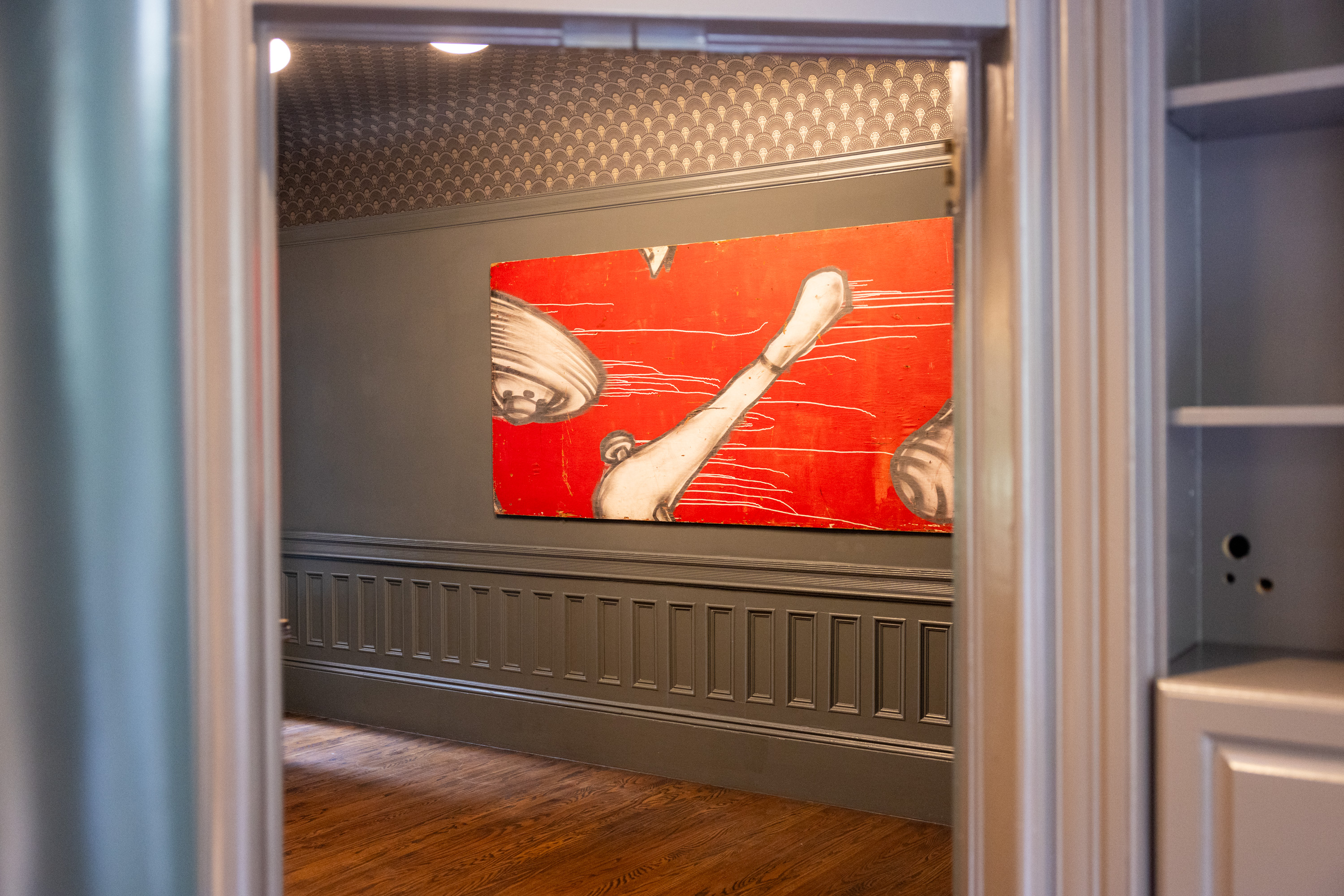 The image shows a room with grey wainscoted walls and wooden flooring, featuring a red abstract painting with white and black shapes, visible through a doorway.