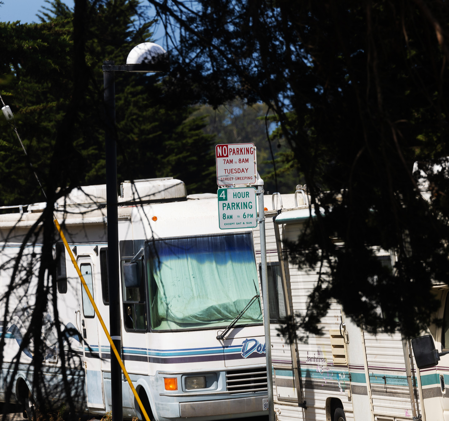 The image shows an RV parked on the street, partially obscured by trees. There are parking restriction signs indicating no parking on Tuesdays from 7-8 AM and a four-hour limit from 8 AM to 6 PM.