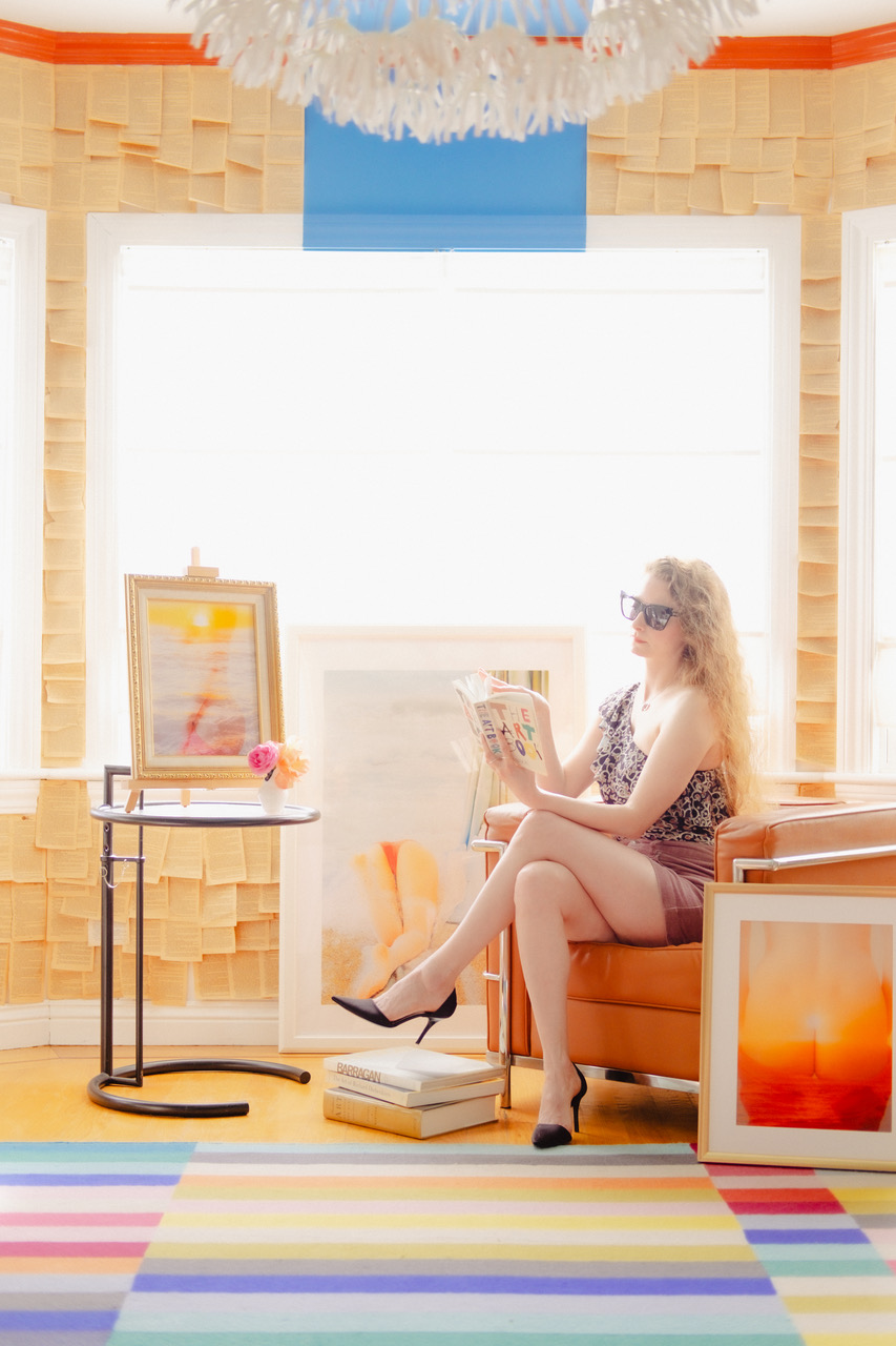 A woman with long hair and sunglasses reads a book while sitting on an orange chair in a brightly lit room, decorated with art and pages on the walls.