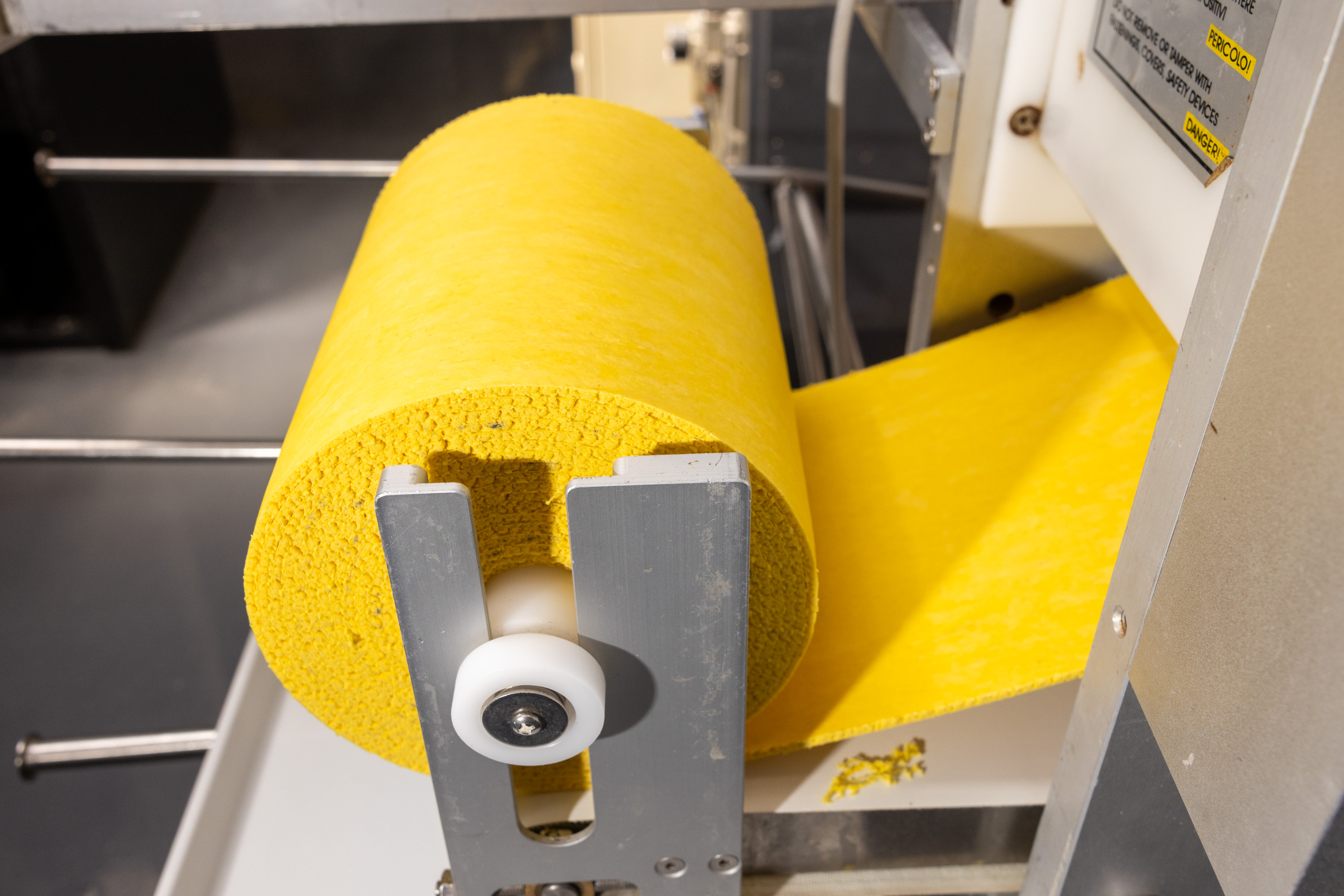 The image shows a large yellow roll of material mounted on a metal dispenser, with the material being fed through the machine, possibly for processing or cutting.