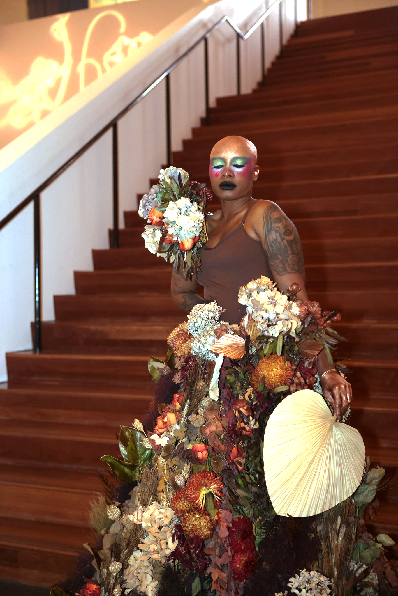 A person with bold, colorful makeup stands on a staircase, wearing a dress adorned with various dried flowers and holding a bouquet and a large leaf fan. They have tattoos.