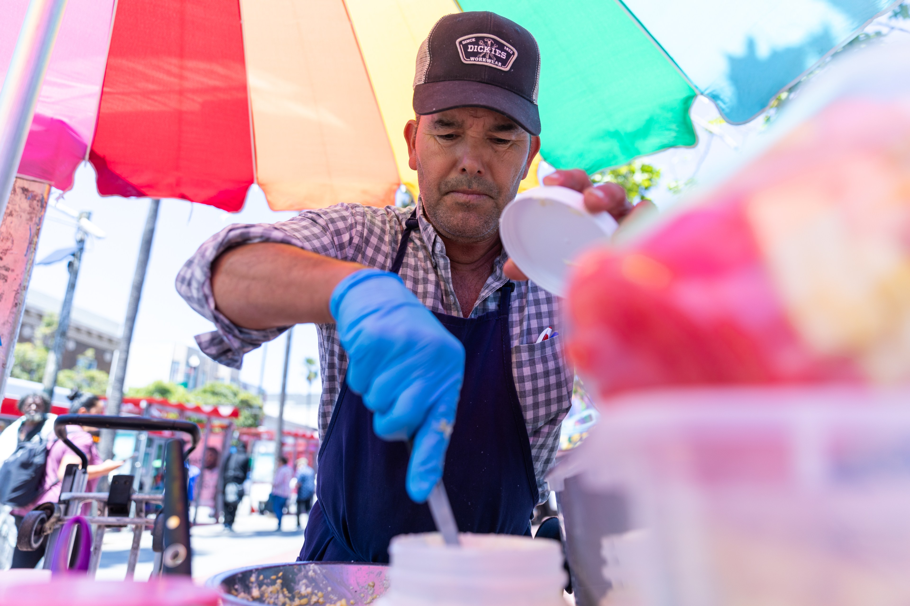 A man wearing a checkered shirt, blue apron, and gloves is preparing food under a brightly colored umbrella at an outdoor market or street fair.
