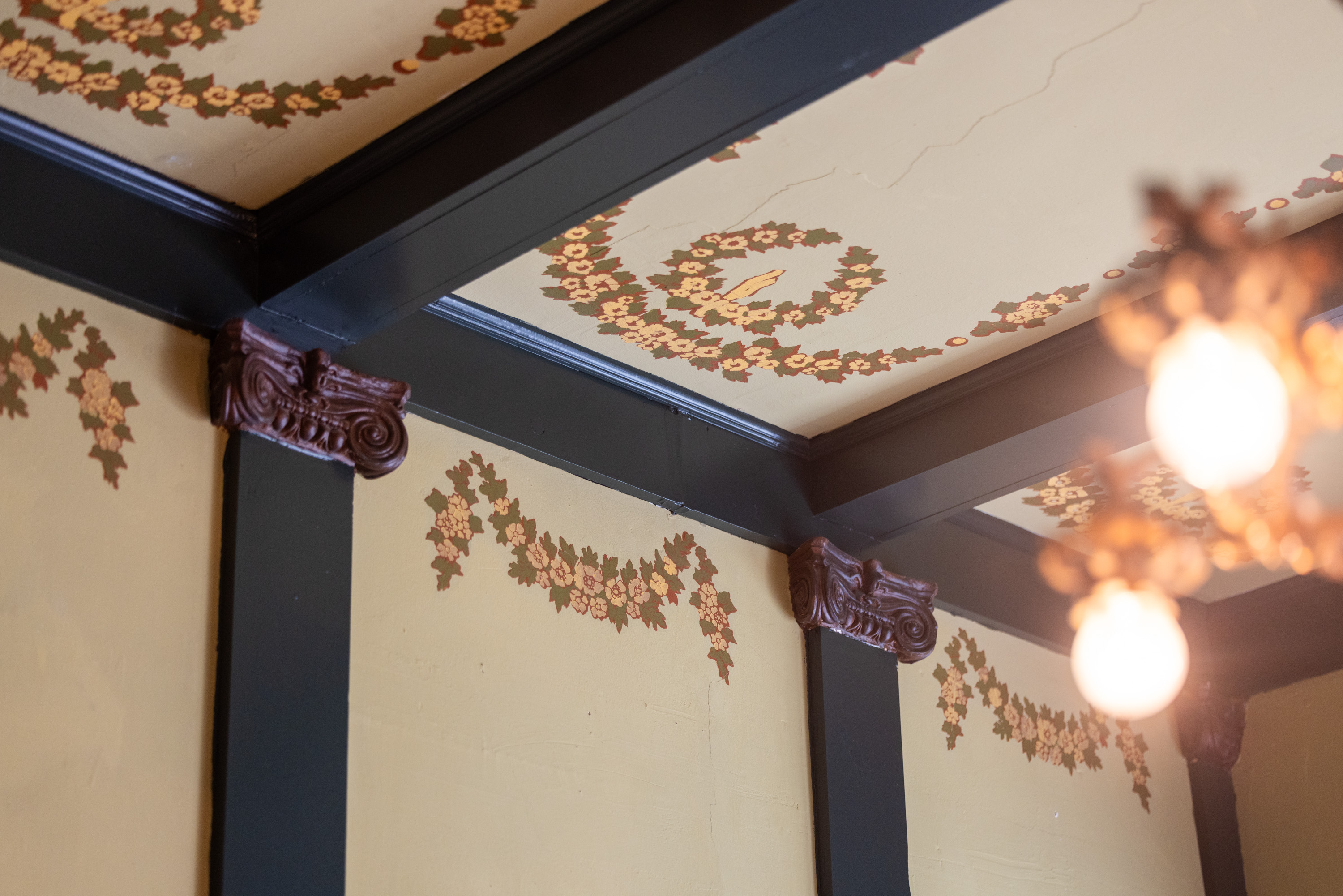 The image shows a room corner with decorative floral paintings on the walls and ceiling, dark wooden beams, ornate corner brackets, and a partially visible chandelier.