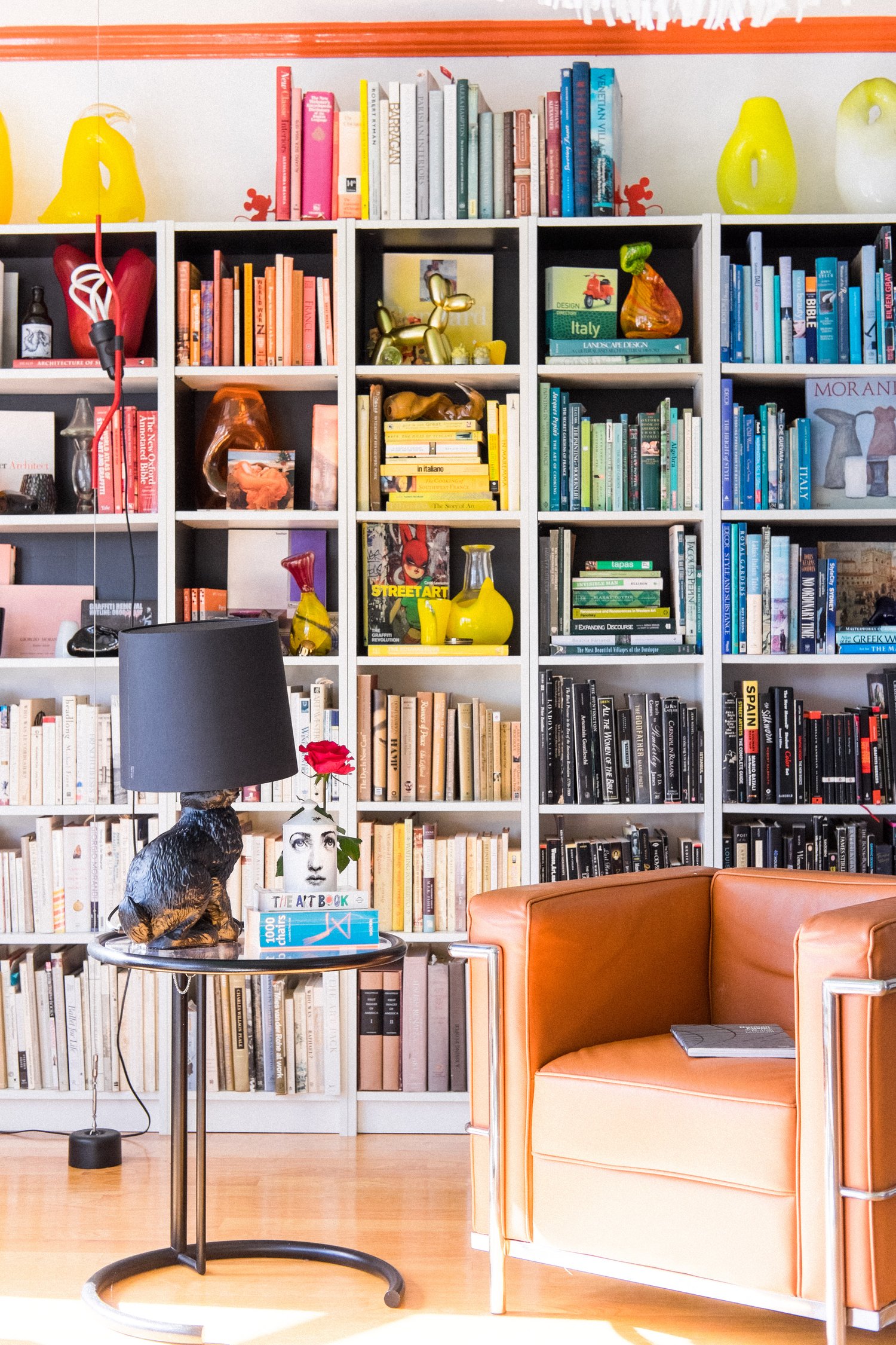 The image shows a colorful bookshelf organized by book color, with various decorative items. In front of the shelf is a brown leather armchair and a side table with books and a lamp.