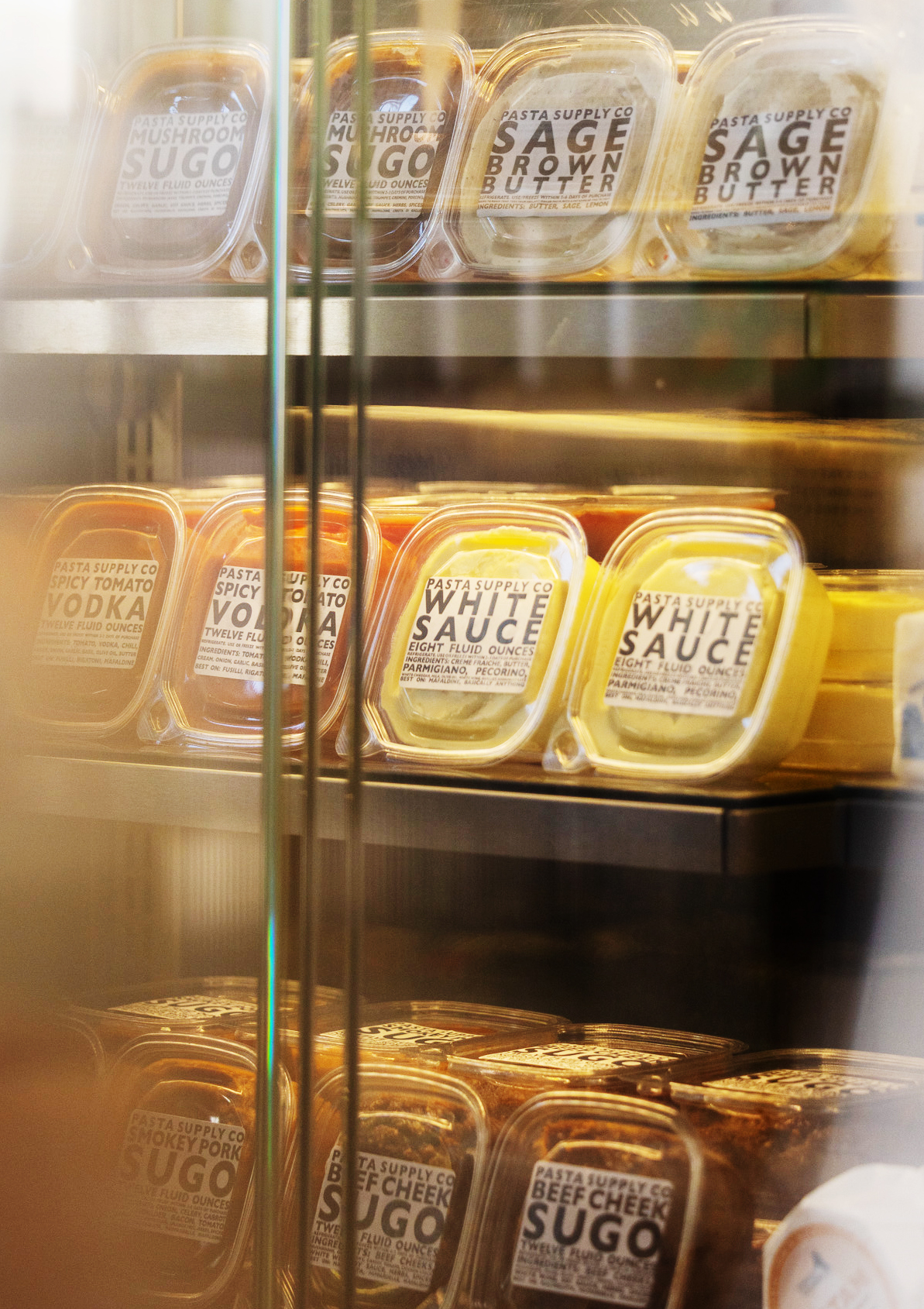 The image shows a display fridge stocked with various pasta sauces in clear plastic containers labeled with flavors like mushroom, sage brown butter, tomato vodka, and white sauce.