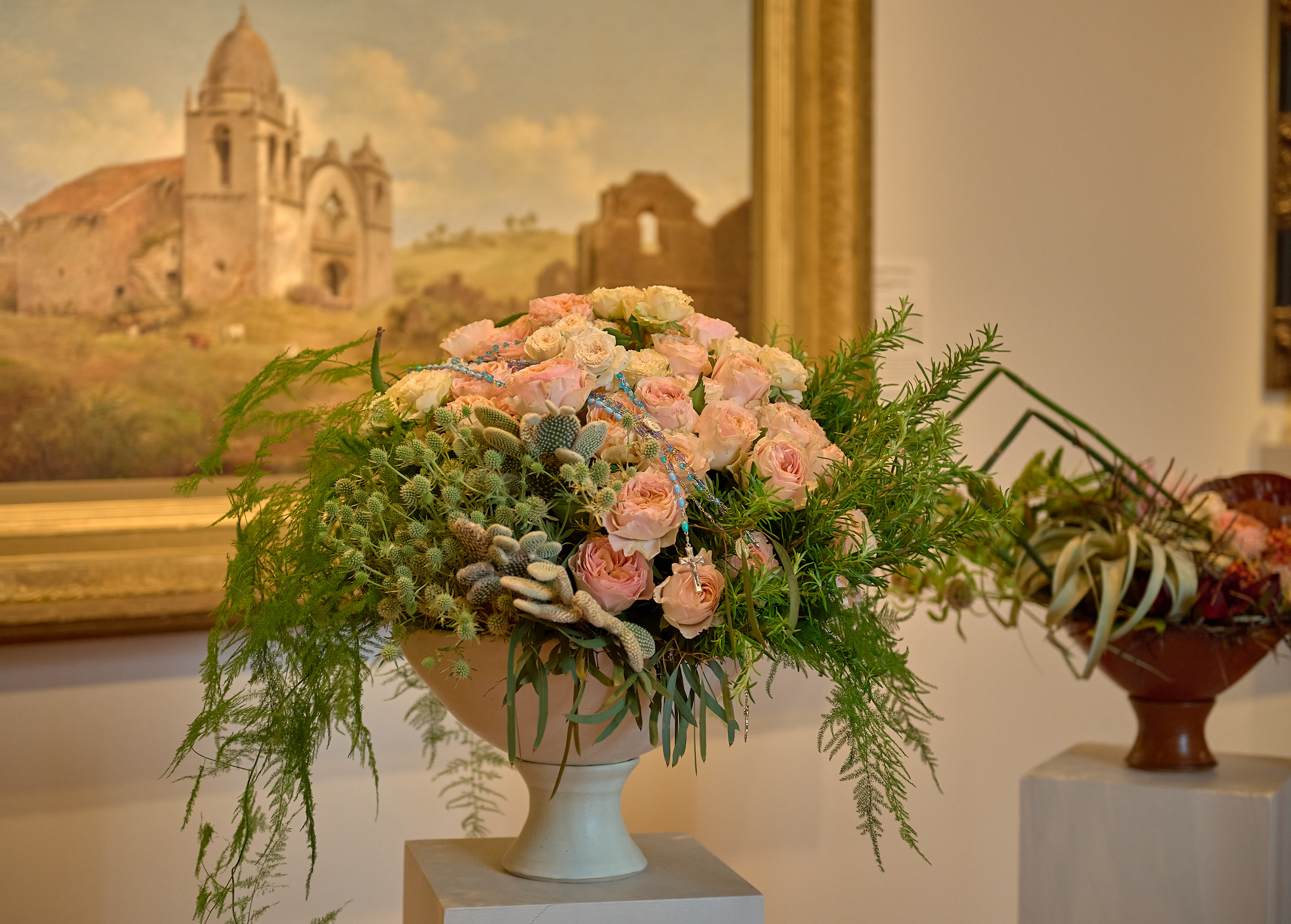 A floral arrangement with pink roses and green foliage in a white pedestal vase is in the foreground. A painting of an old building is in the background.