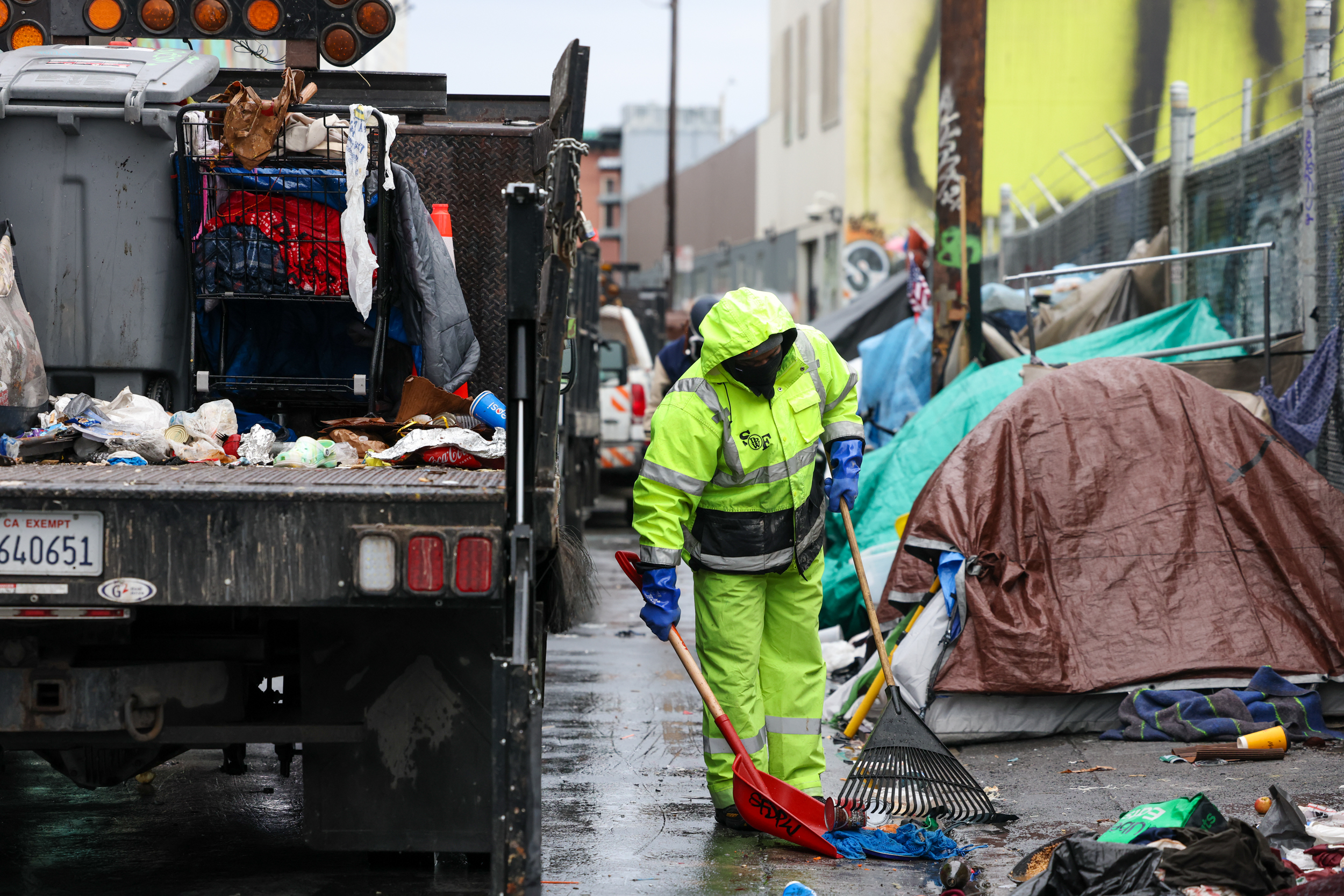 A worker in a bright yellow rain suit uses a shovel and rake to clean a trash-littered street beside a sanitation truck and a makeshift tent encampment.