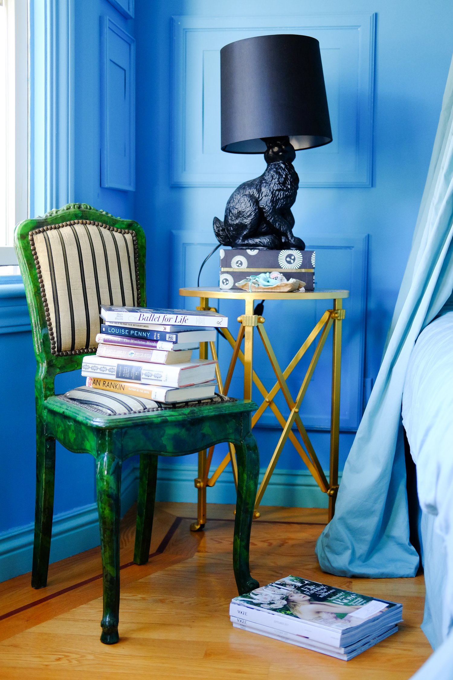 The image shows a blue-painted room with a green chair, a pile of books on it, a small gold side table with a black animal-shaped lamp, and magazines on the wooden floor.