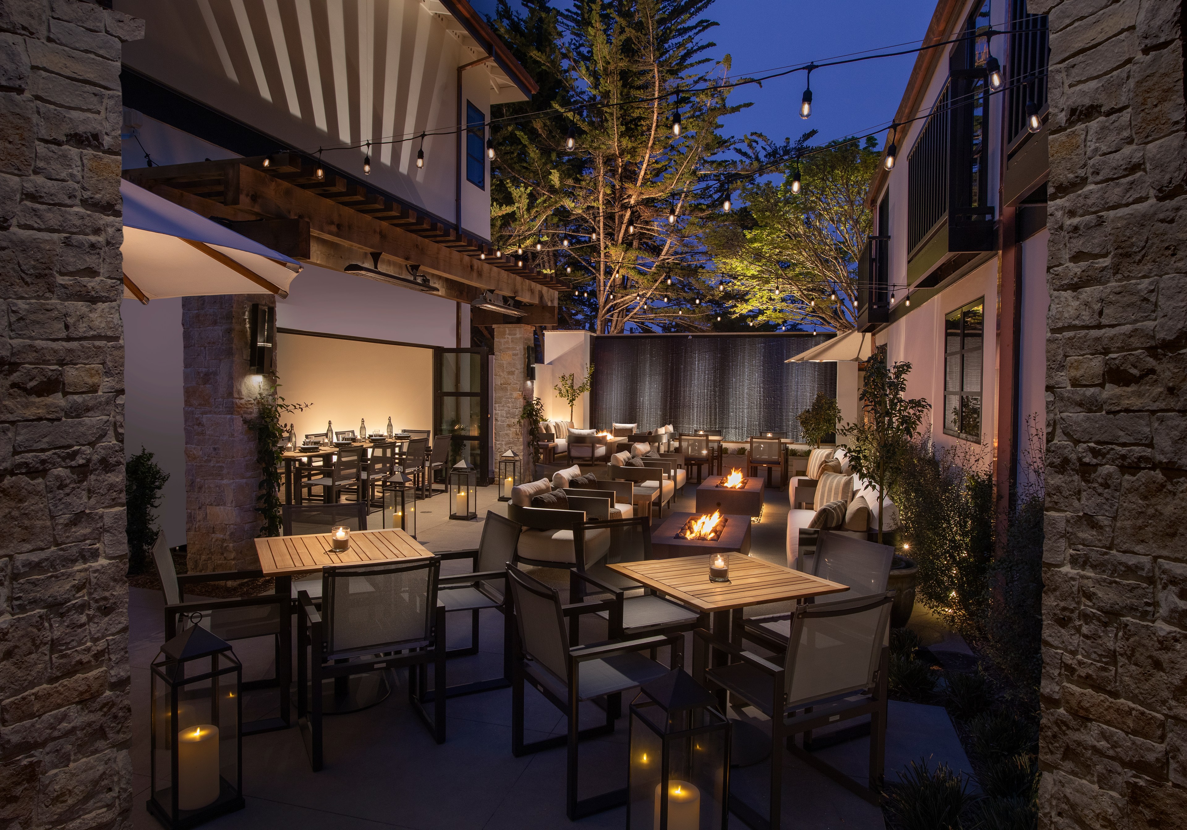 An enchanting outdoor patio scene at night is illuminated by string lights, with cozy seating, tables set with candles, and warm fire pits. Trees frame the background.