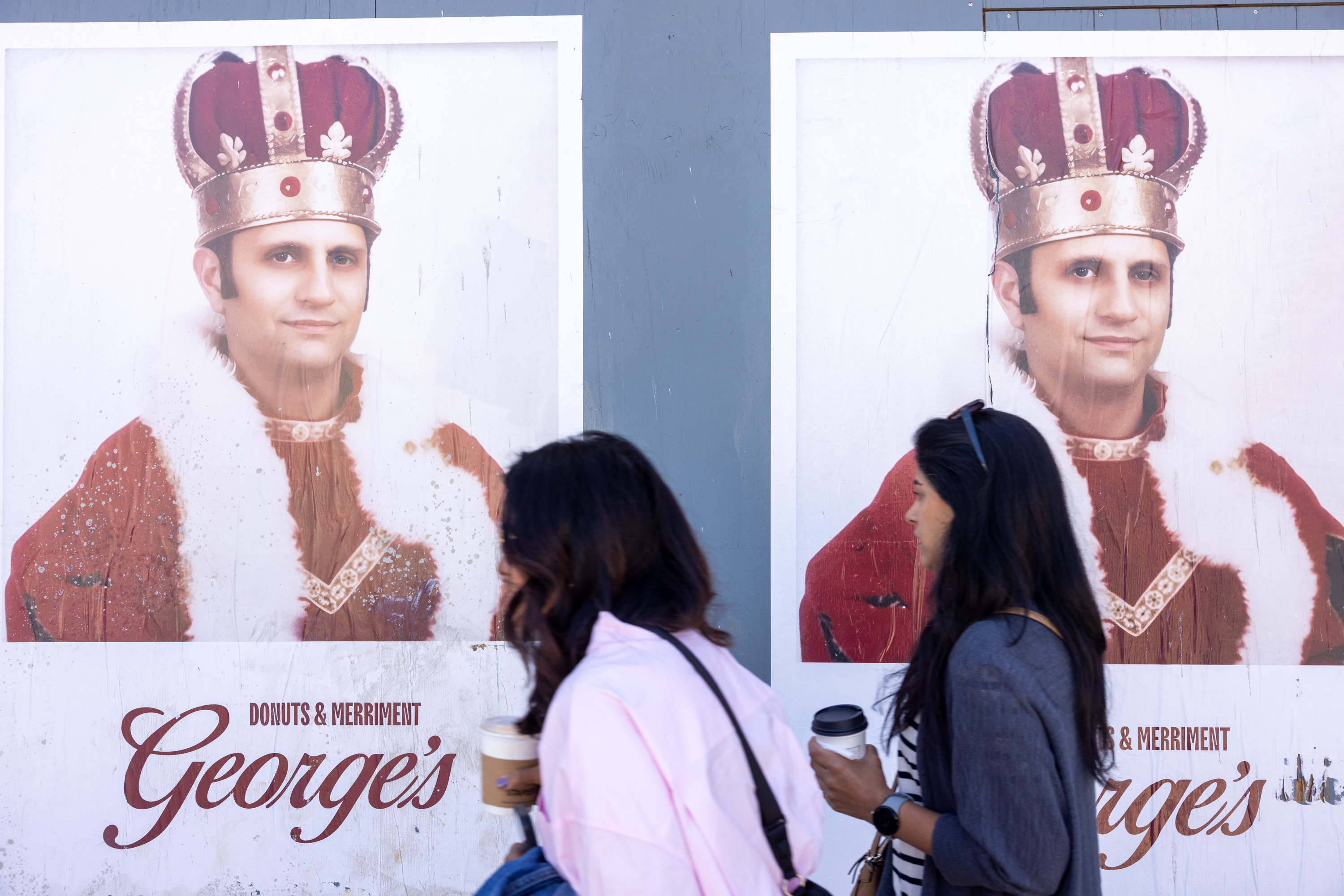 The image shows two women walking past posters of a man dressed as a king, advertising George's Donuts &amp; Merriment. Each woman is holding a drink in her hand.
