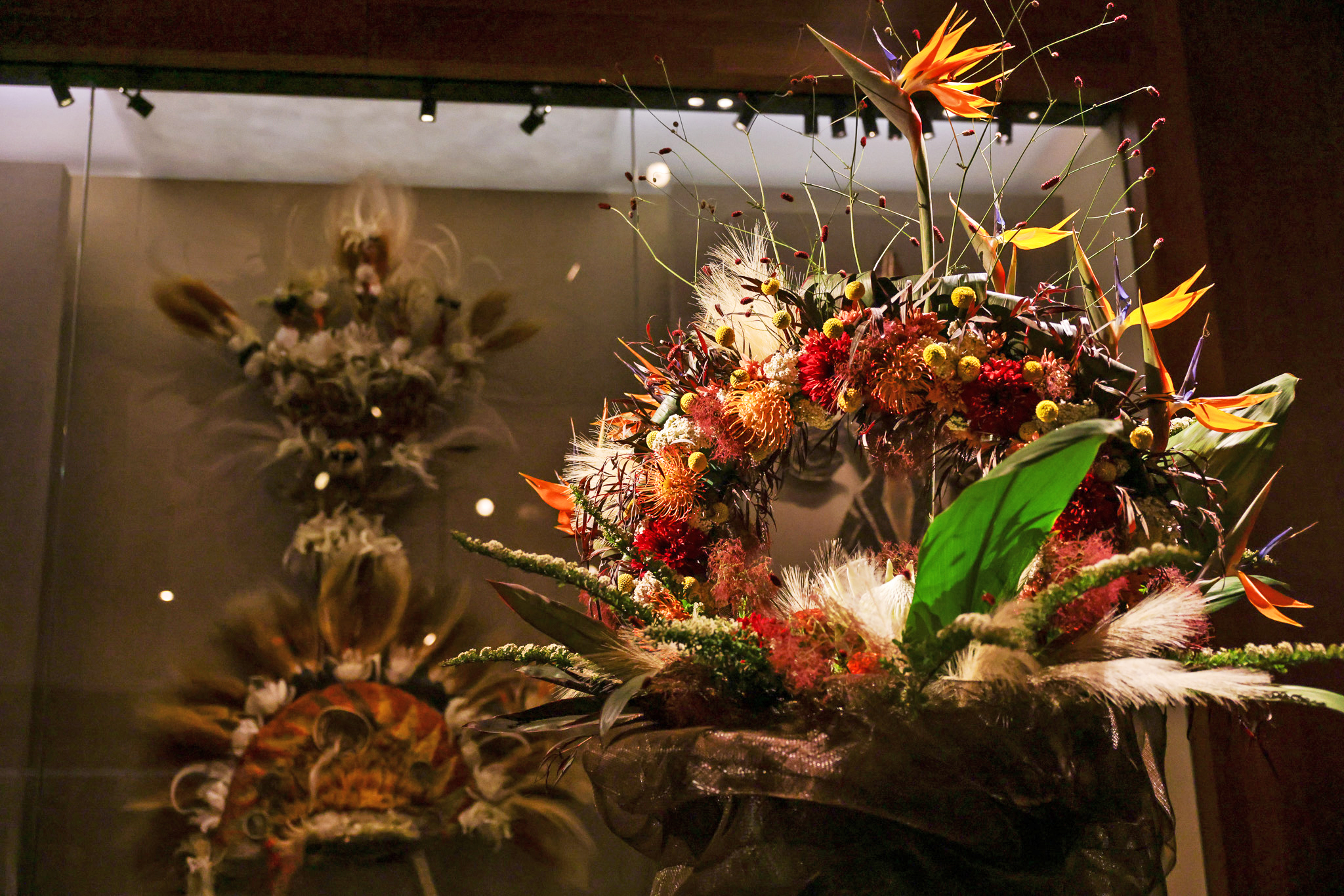 The image features an ornate floral arrangement with vibrant orange, red, and yellow flowers, green leaves, and spiky accents, displayed in a museum-like setting.