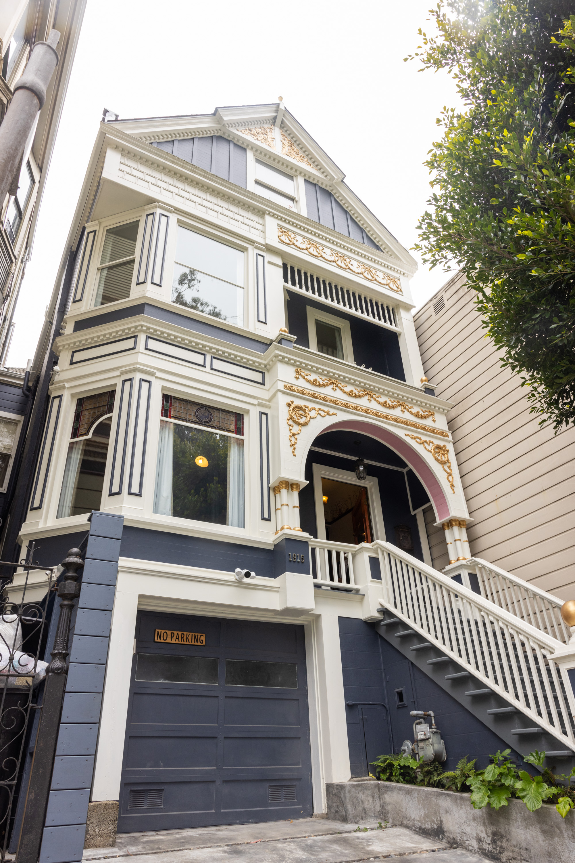 The image shows a multi-story Victorian-style house with ornate gold trim, a bay window, a gray garage labeled &quot;No Parking,&quot; and an external staircase.