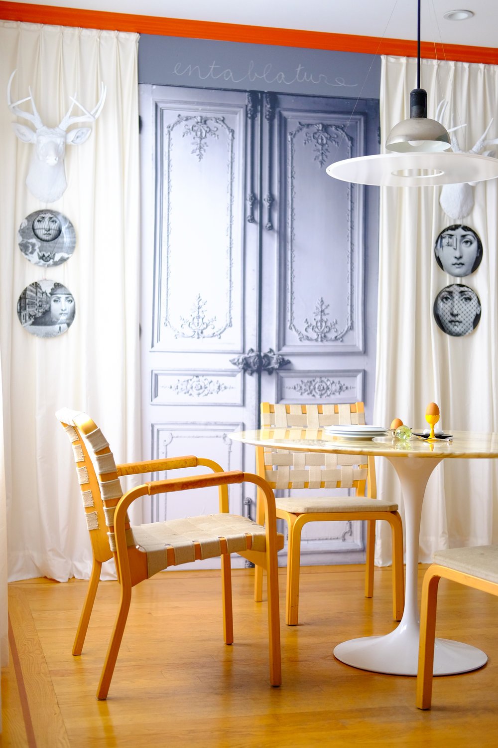 A dining area with a round white table, light wooden chairs, and a pendant light. The backdrop features ornate double doors, accented by plates and deer head wall decor.