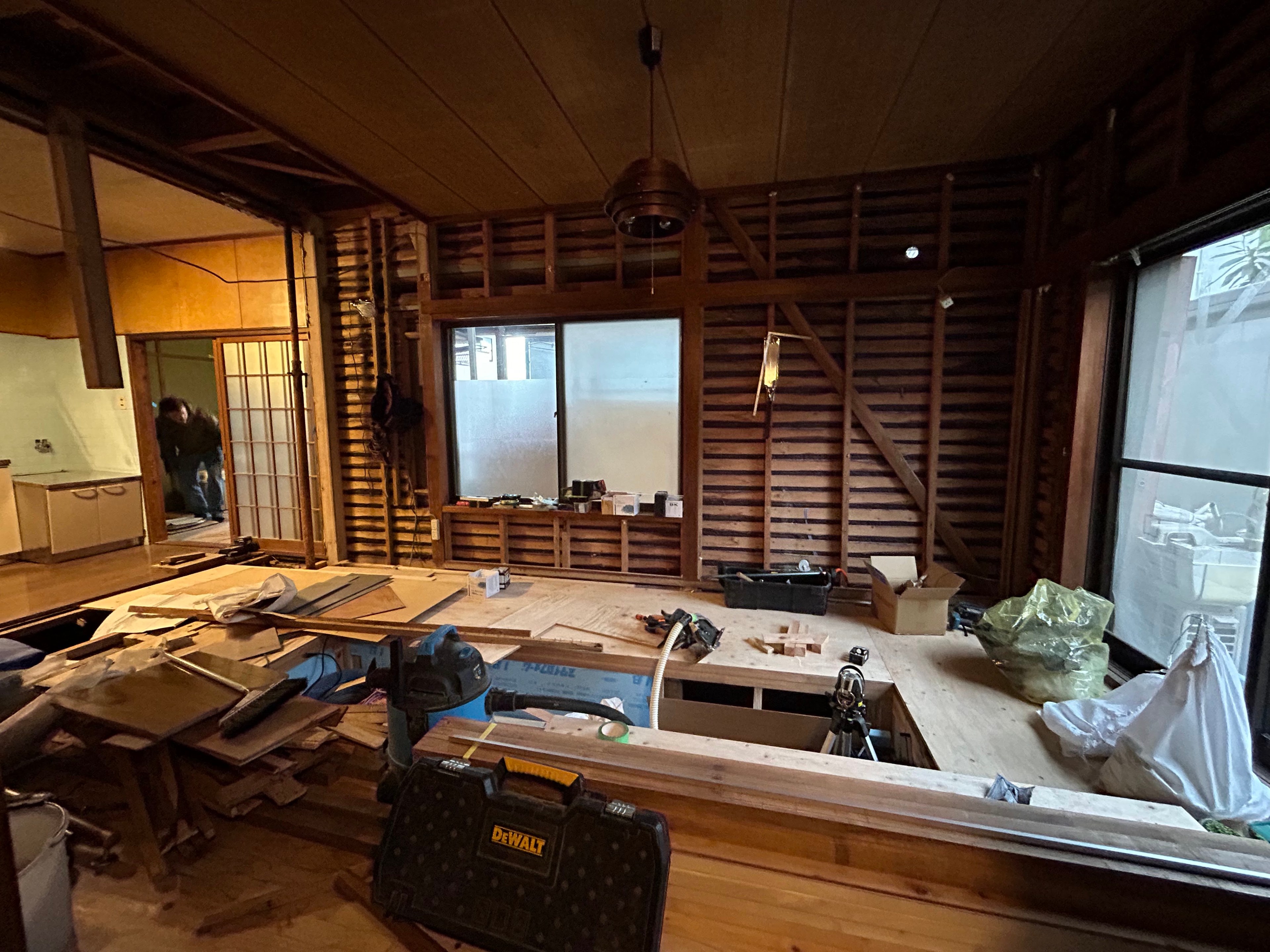 The image shows a room under renovation with exposed wooden beams, construction tools, and materials scattered around. There are large windows and a sliding door.