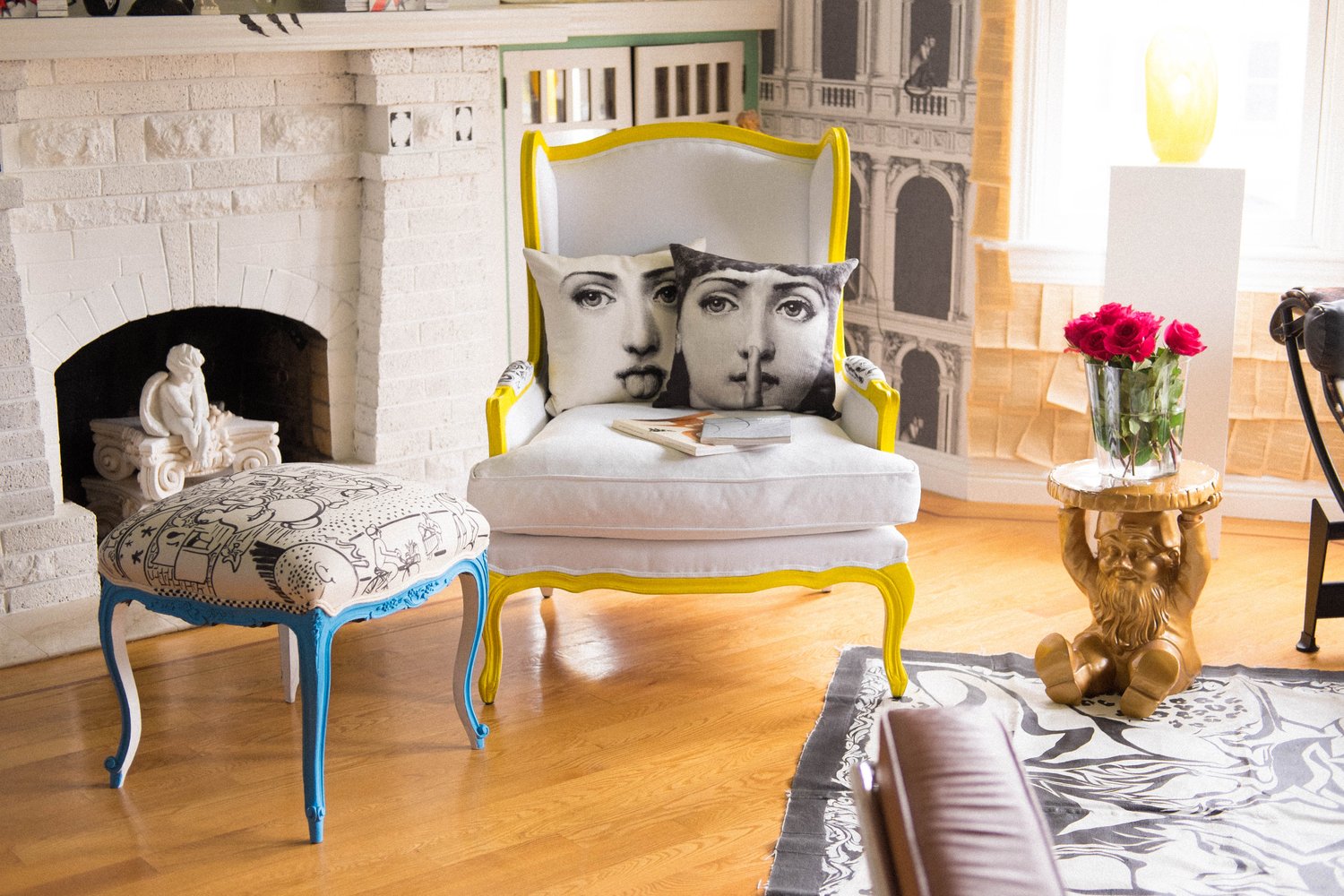 The image shows a cozy living area featuring a white armchair with yellow trim, two pillows with faces on them, and a small table, supported by a golden sculpted figure holding a vase of red roses.