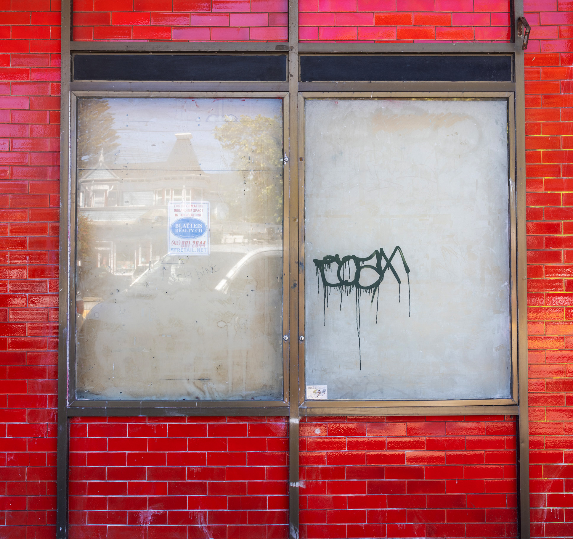 The image shows a red brick wall with two large windows. One window has a &quot;for sale&quot; sign and the other has black graffiti. The reflections show building facades.