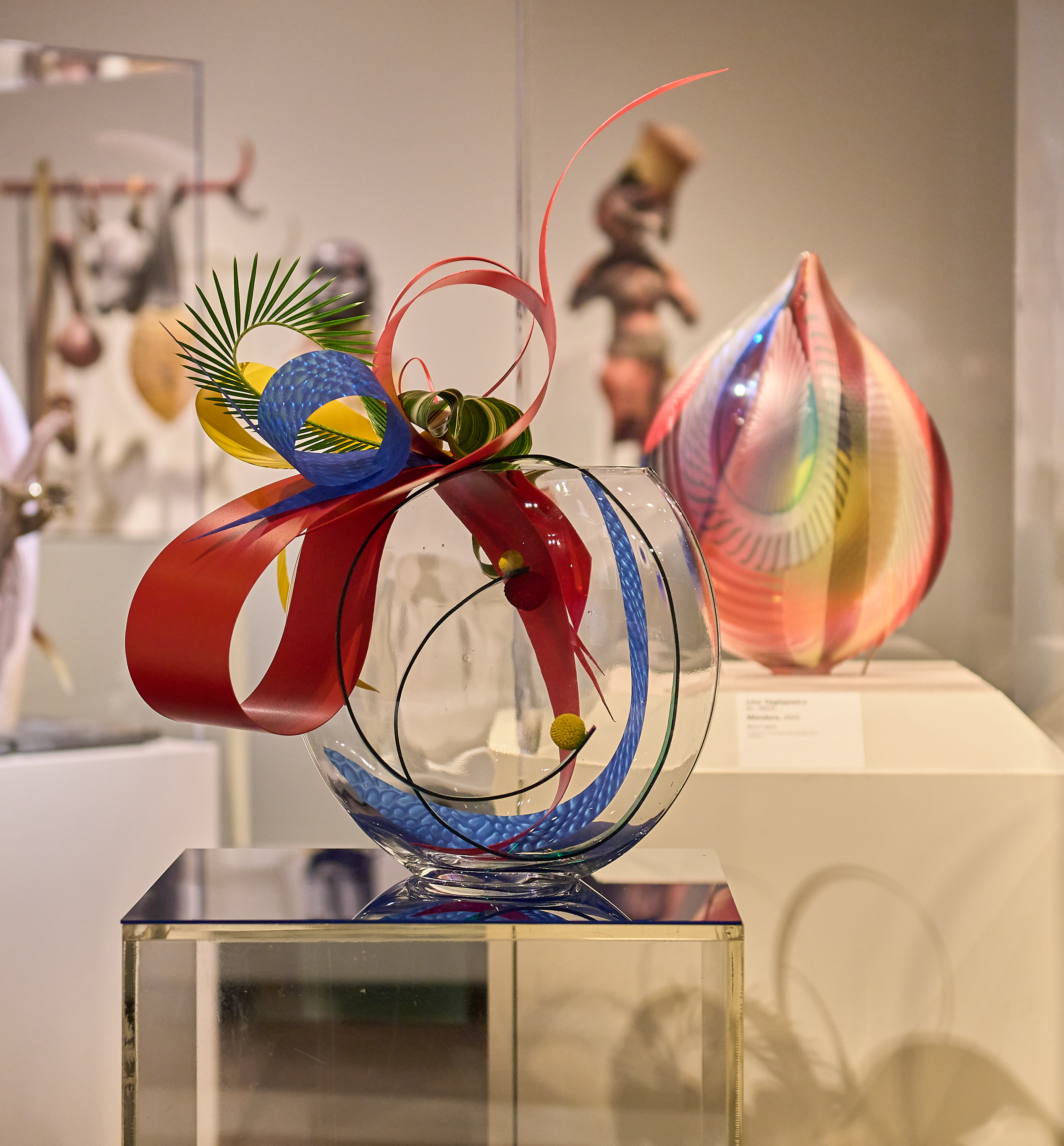 A glass vase with intricate designs, featuring red, blue, and yellow decorations, sits on a pedestal, with colorful abstract art pieces displayed in the background.