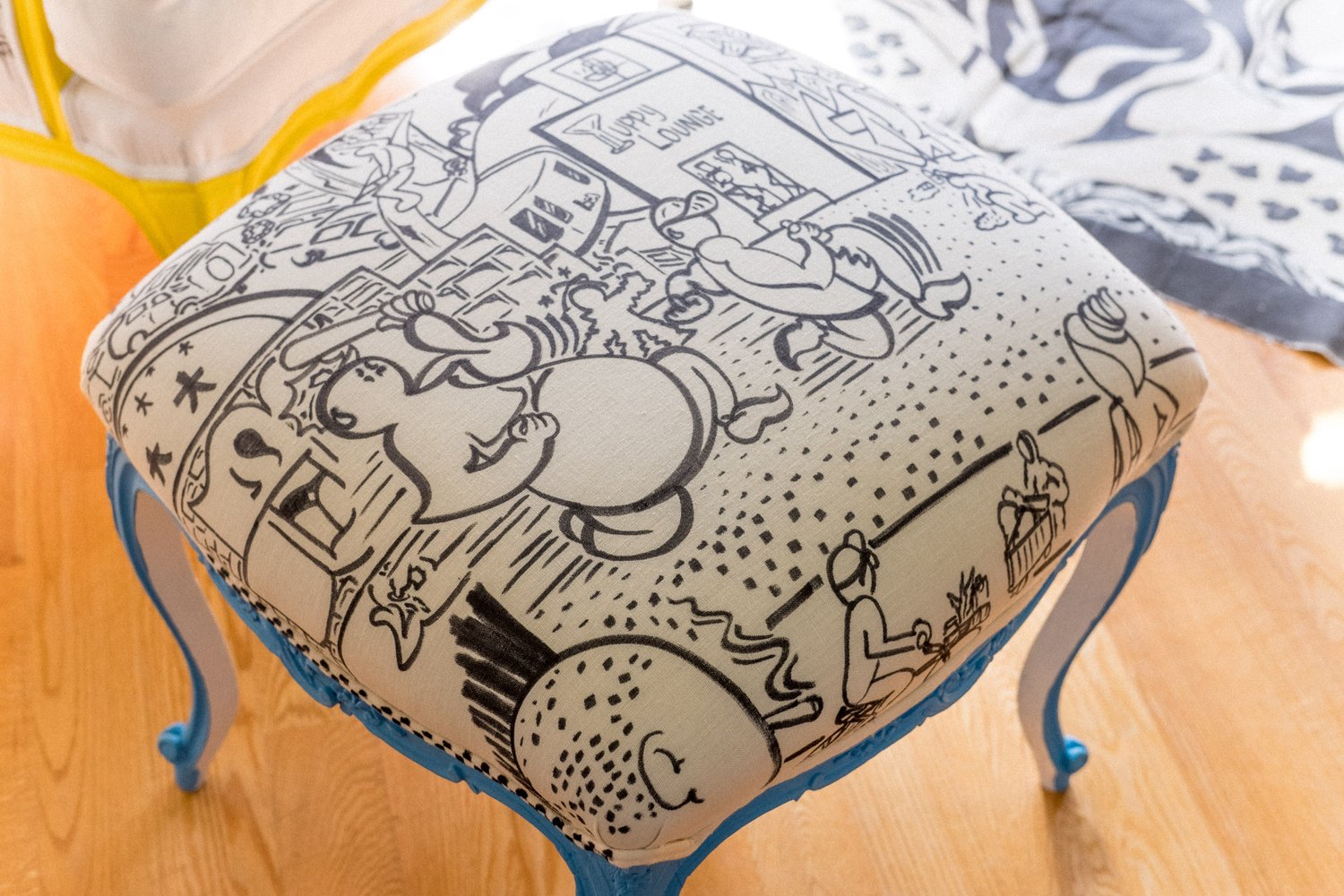 The image shows a stool with blue legs featuring a cushion adorned with whimsical black-and-white cartoons, including various characters and scenes in an urban setting.