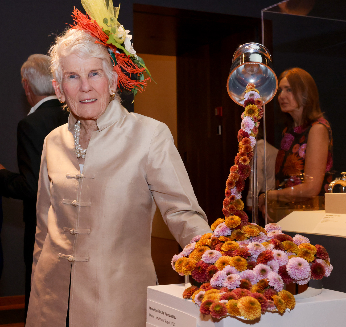 An elderly woman with white hair and a vibrant, floral hat stands next to a decorative arrangement of multicolored flowers that appears to be cascading from a fixture.