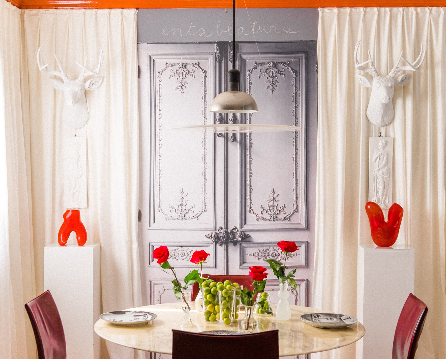 A chic dining area features a round table with roses, green grapes, ornate gray doors, white curtains, deer busts, modern red sculptures, and a hanging lamp.