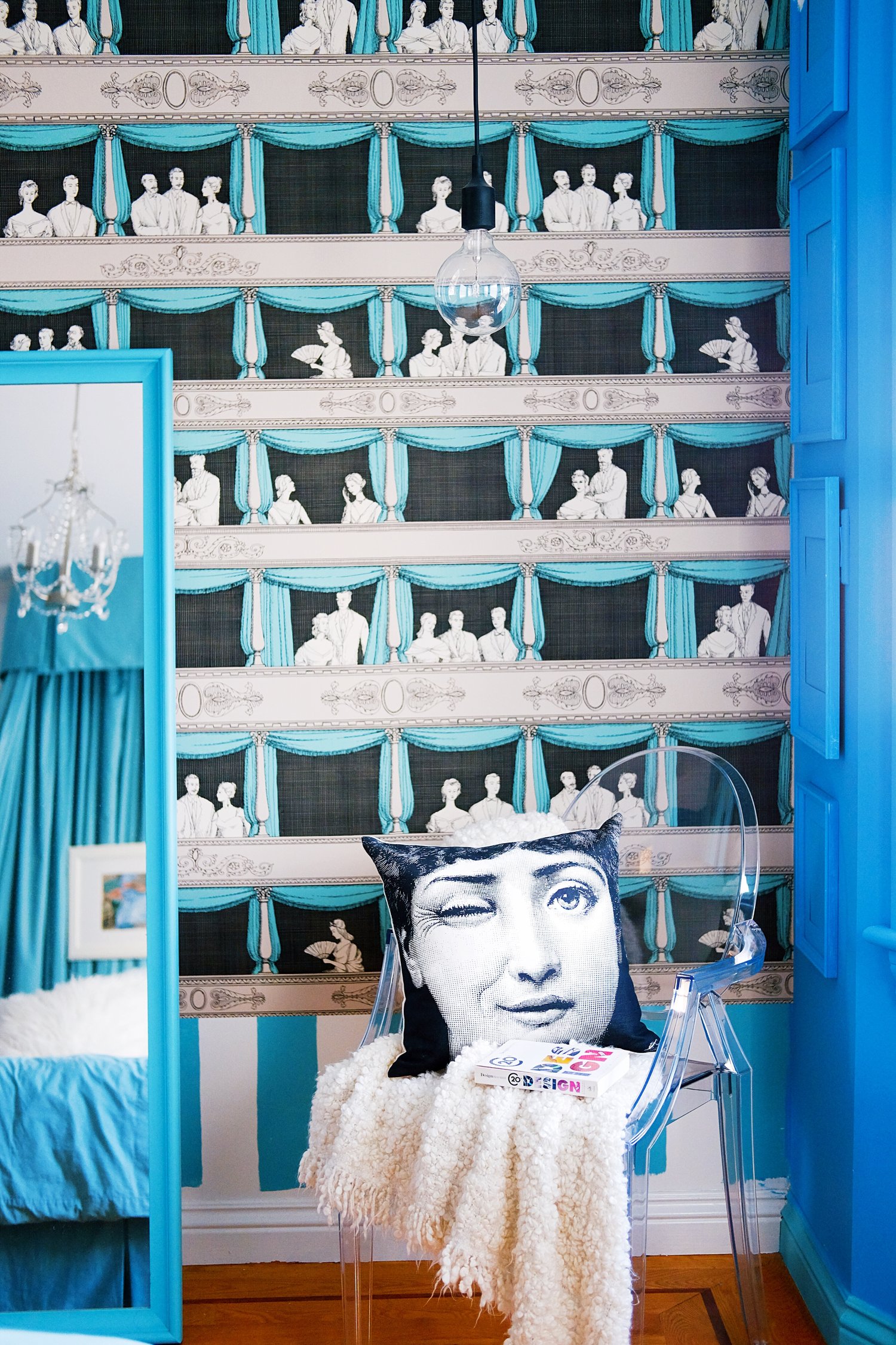 The room features whimsical wallpaper depicting vintage theater scenes, a clear chair with a winking face pillow and books, a blue-framed mirror, and a hanging bulb.