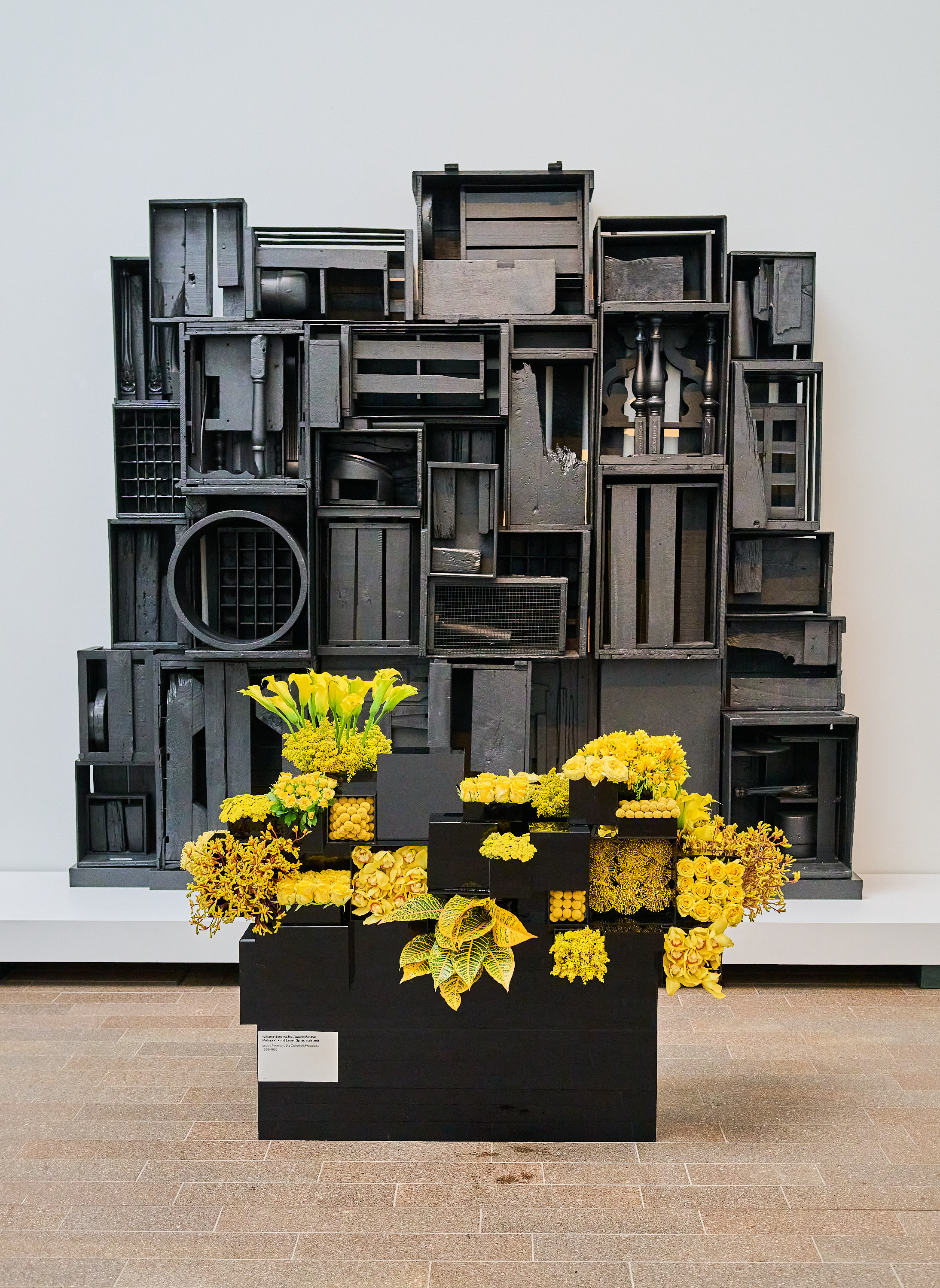 The image shows an abstract art installation with stacked black boxes and compartments filled with assorted yellow flowers and foliage, displayed against a white wall.