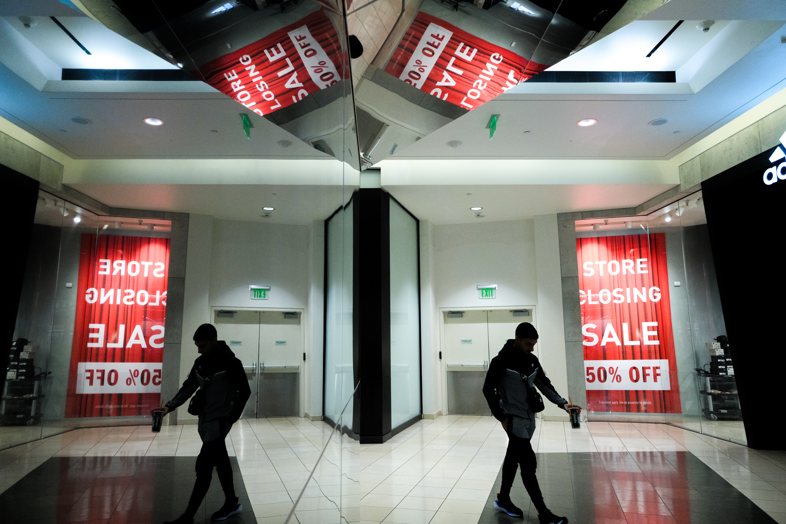 A person walks past mirrored walls in a mall, holding a drink. Large red signs read &quot;STORE CLOSING SALE 50% OFF&quot; in the background.