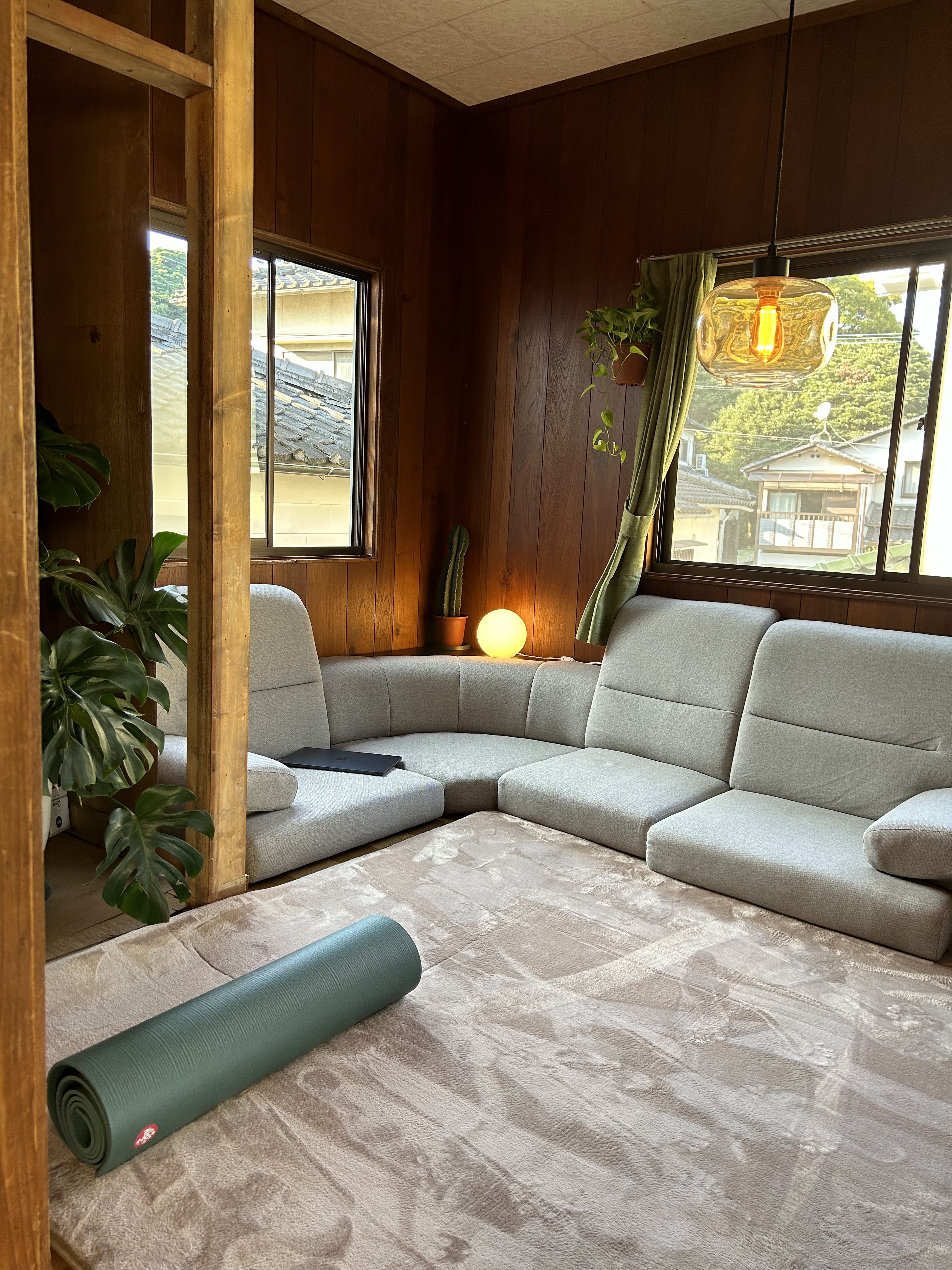 The image shows a cozy room with wood-paneled walls, featuring a grey L-shaped sofa, a round glowing lamp, a hanging plant, and a yoga mat on the floor.