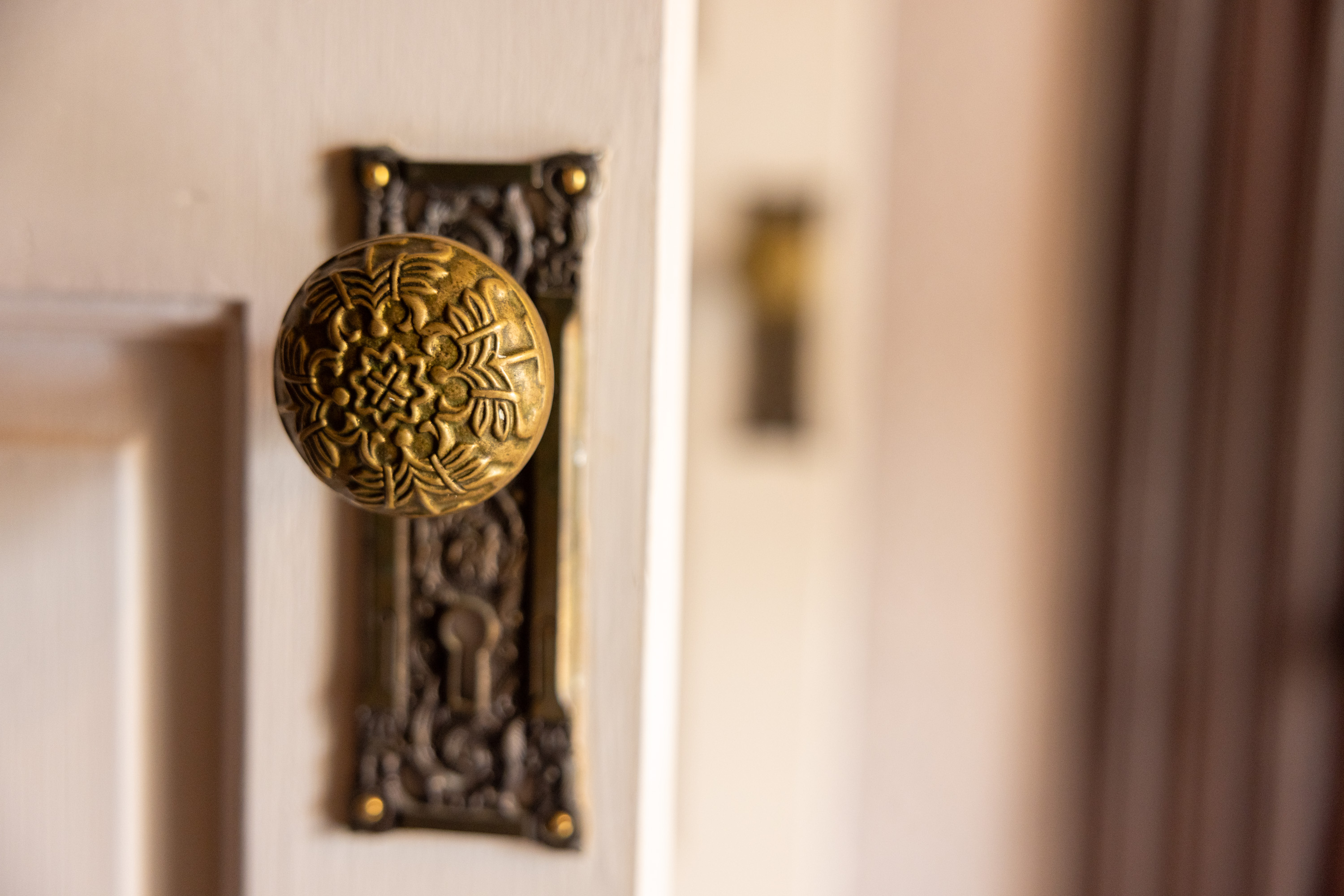 The image shows a close-up of an ornate brass doorknob with intricate floral designs, attached to a detailed brass plate on a white wooden door.