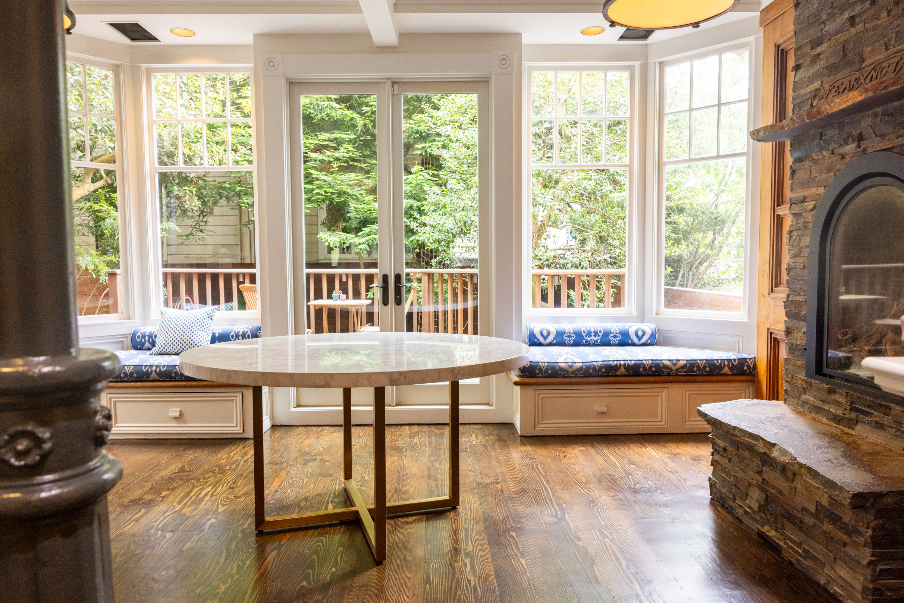 The bright room features a round table, built-in window seats with blue cushions, large windows with greenery outside, and a stone fireplace.