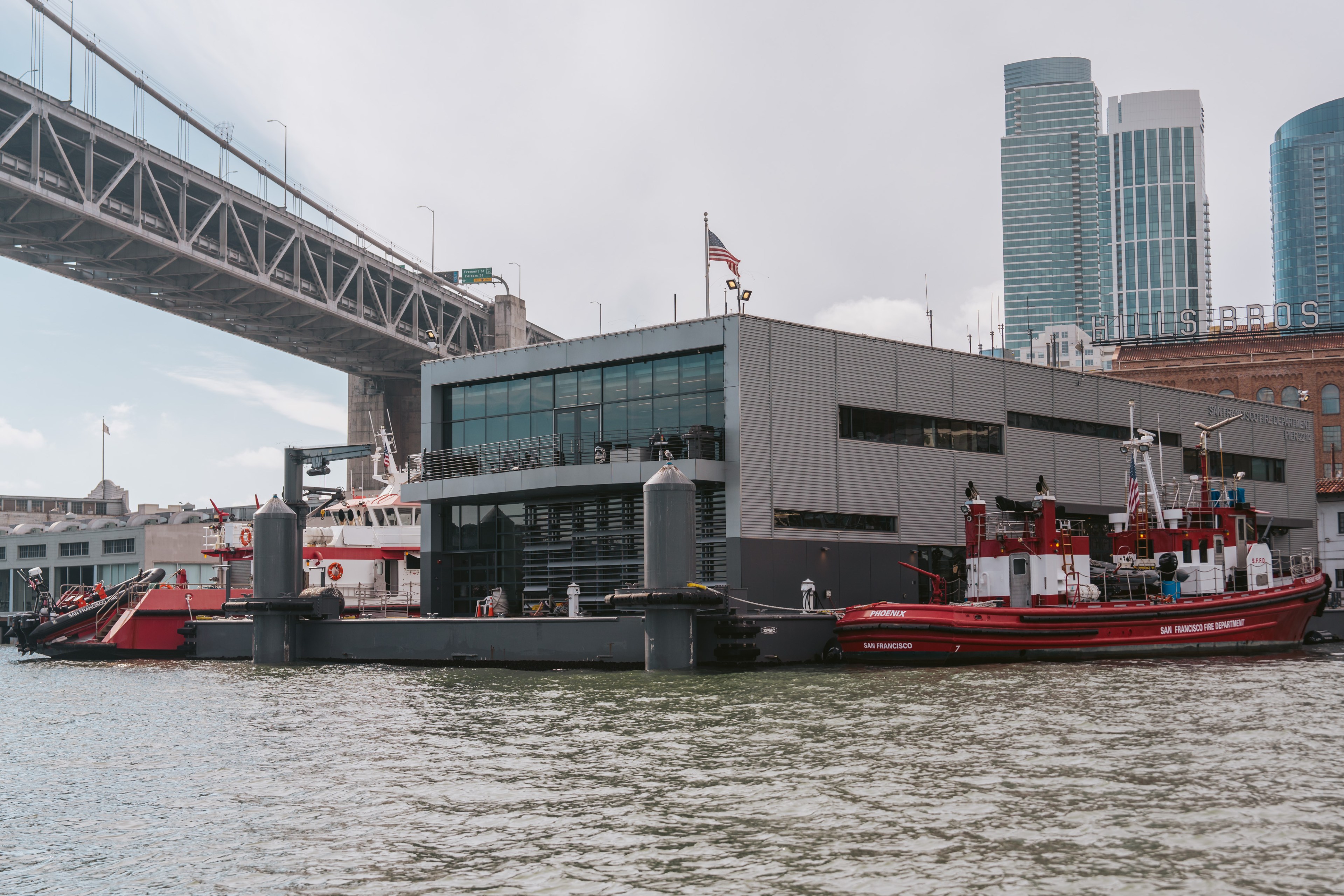 The image shows a modern fire station by the water with a bridge above, high-rise buildings in the background, and fireboats docked at the station.