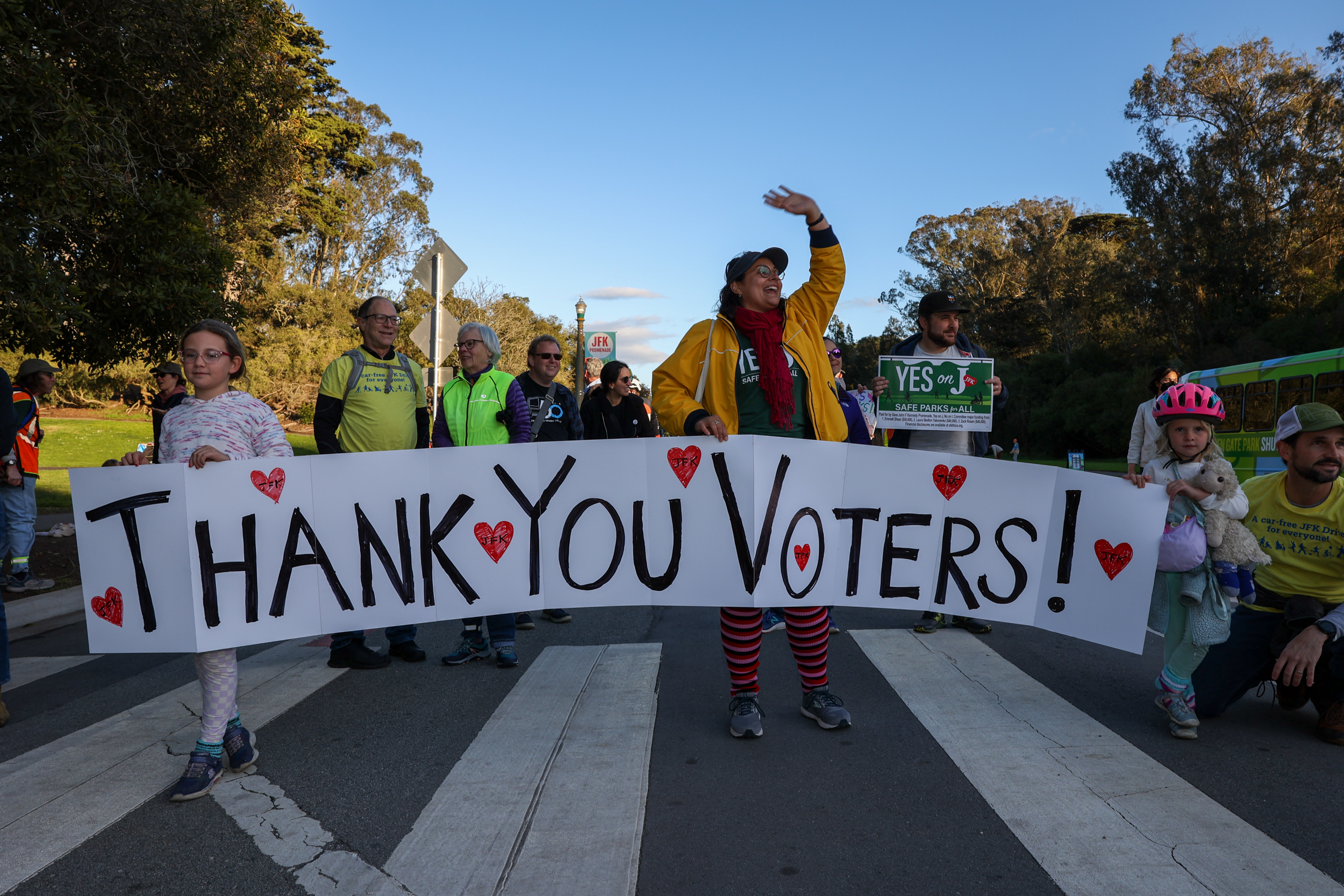 A group of people stand on a street holding a large sign that reads &quot;THANK YOU VOTERS!&quot; with hearts. The scene is outdoors with trees and a clear sky.