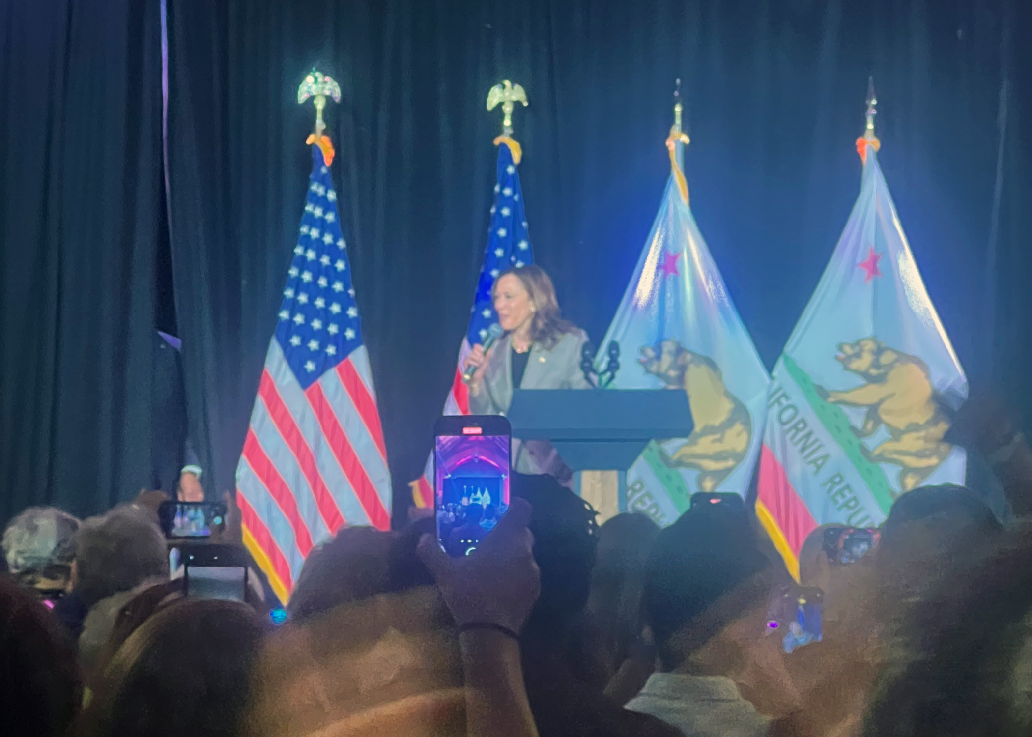 A speaker stands at a podium on a stage, with American and California Republic flags behind them. The audience is holding up phones, capturing the moment.