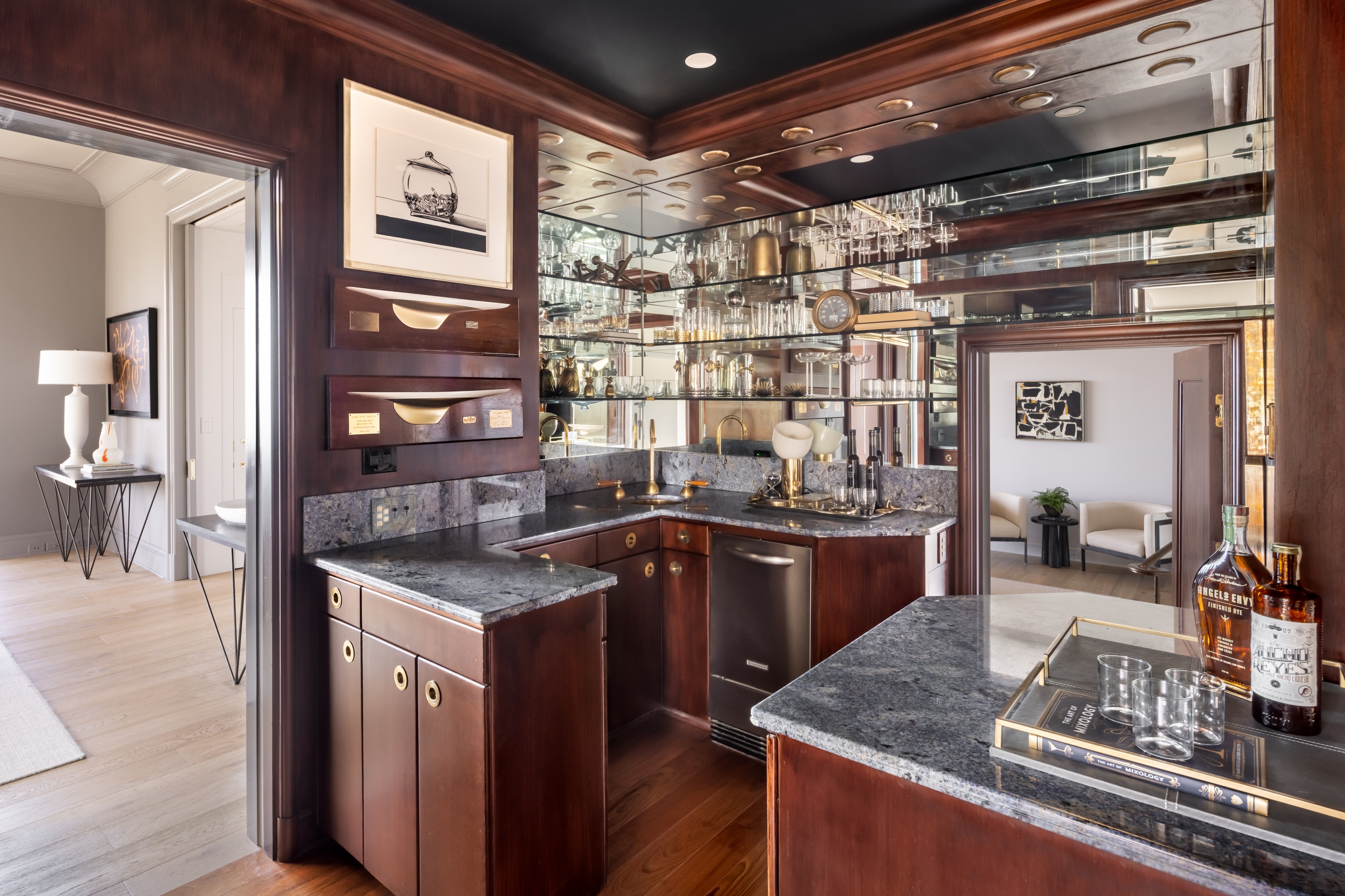 This image depicts an elegant, wood-paneled home bar with granite countertops, mirrored shelving filled with glassware, and modern decor leading to a bright living area.