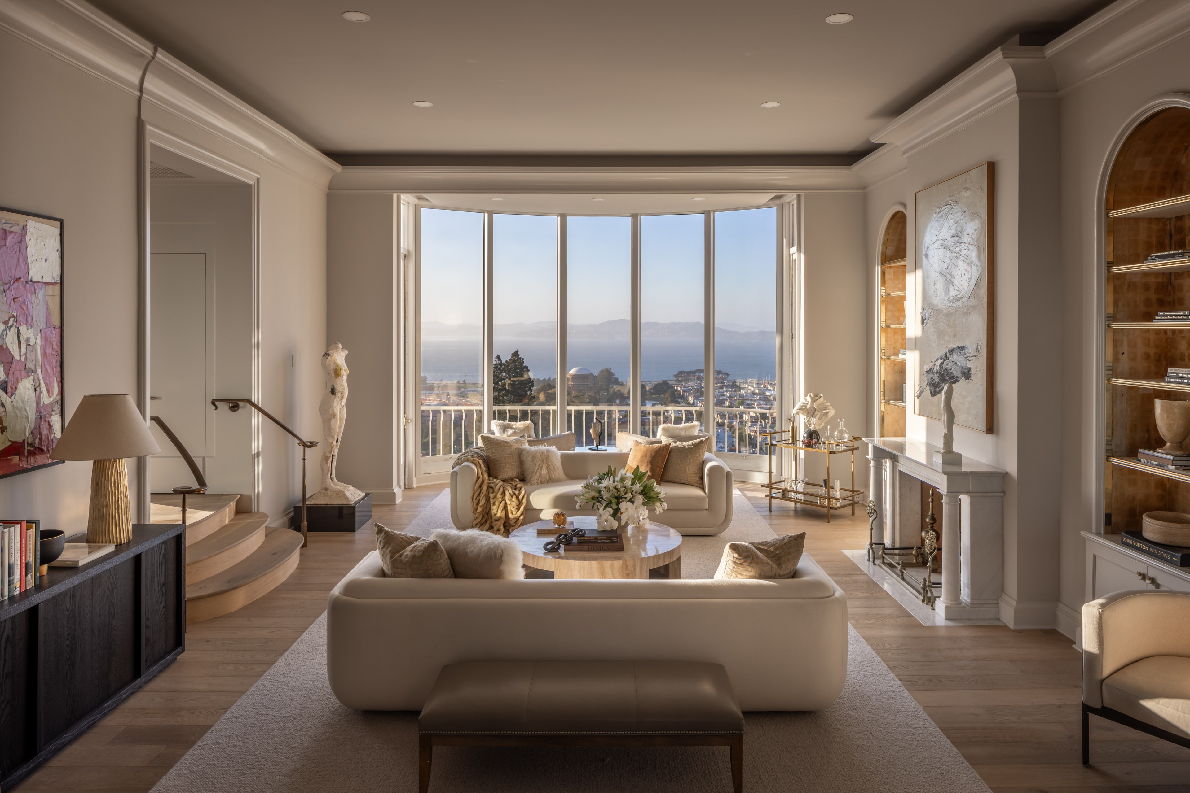 A sophisticated living room with plush white sofas, marble fireplace, large windows offering a coastal view, modern artwork, bookshelves, and elegant decor.